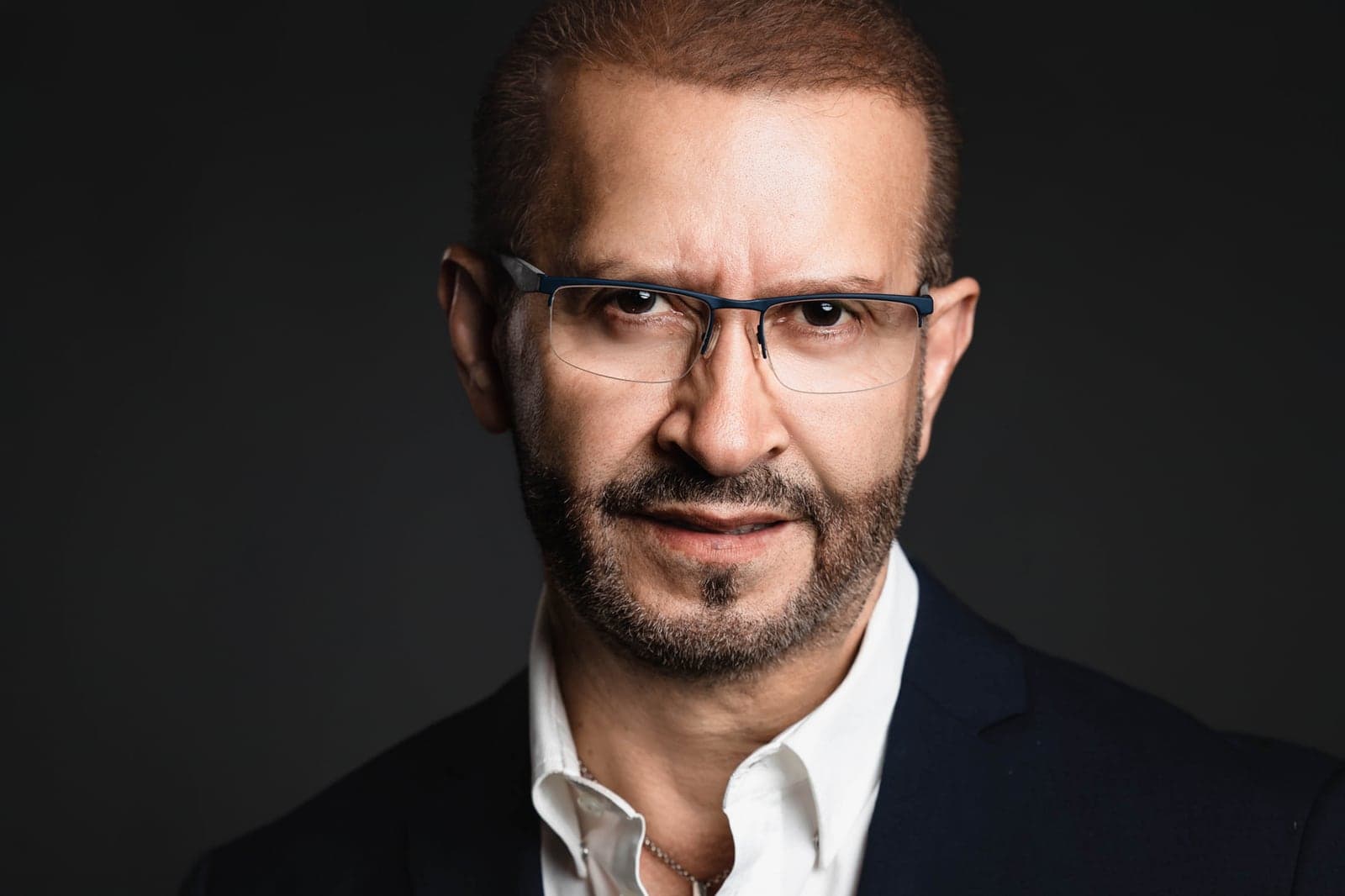 Executive headshot of a man in rimless glasses and a navy blazer on a black background
