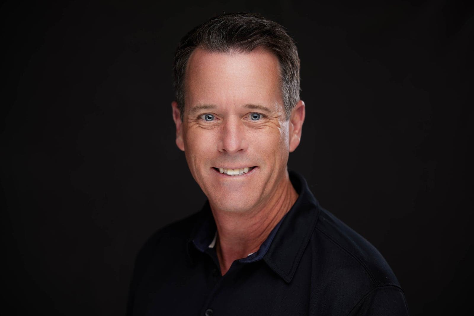 Professional headshot of a smiling man in a black polo on a dark background