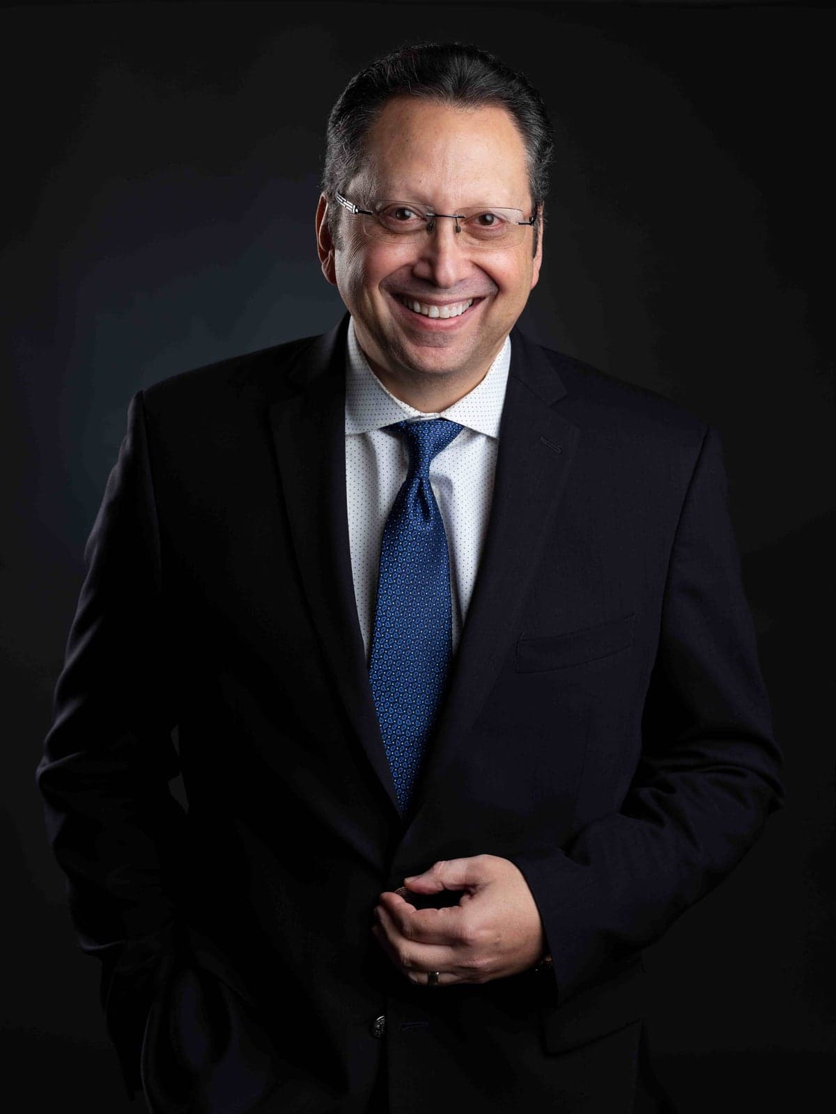 Executive headshot of a man in a navy suit and blue tie on a dark background