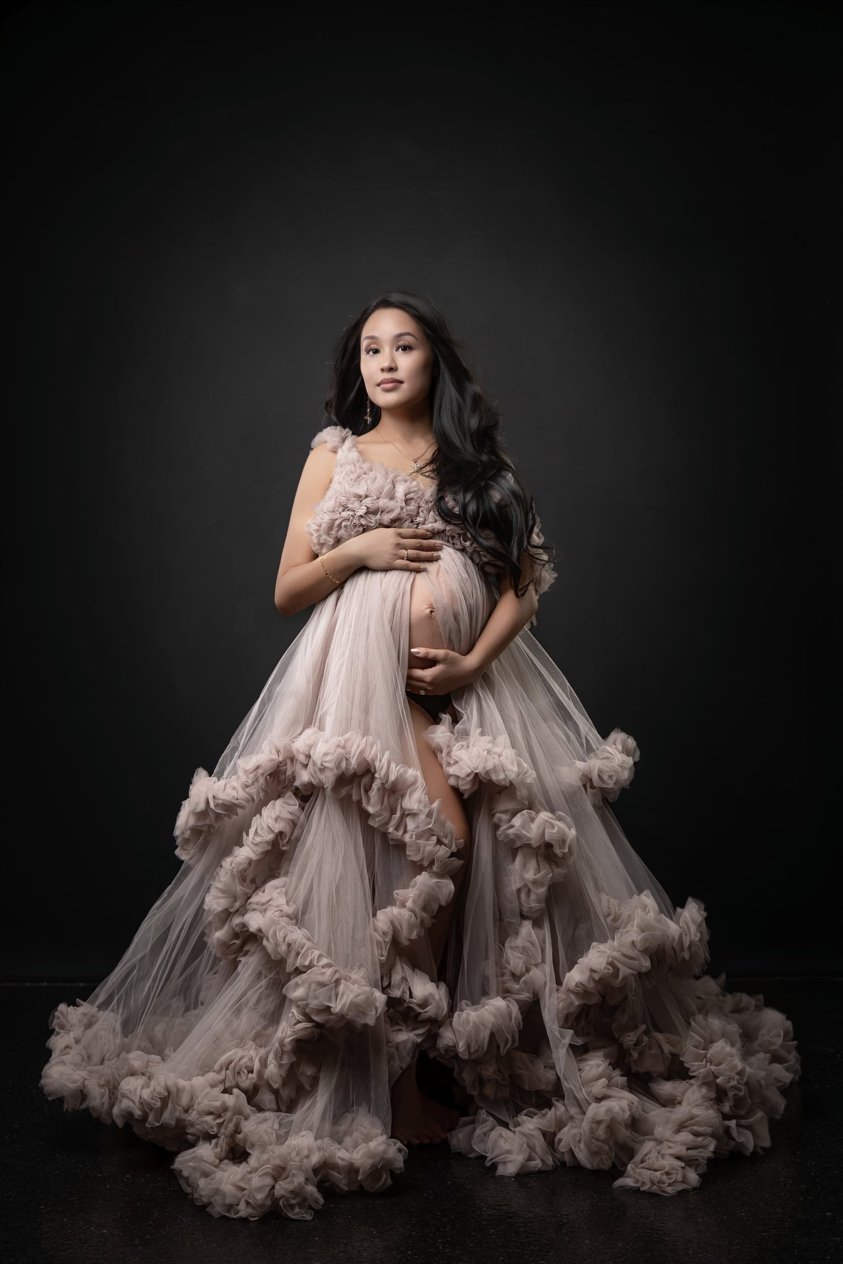 Maternity portrait in a blush ruffled tulle gown on a dark backdrop