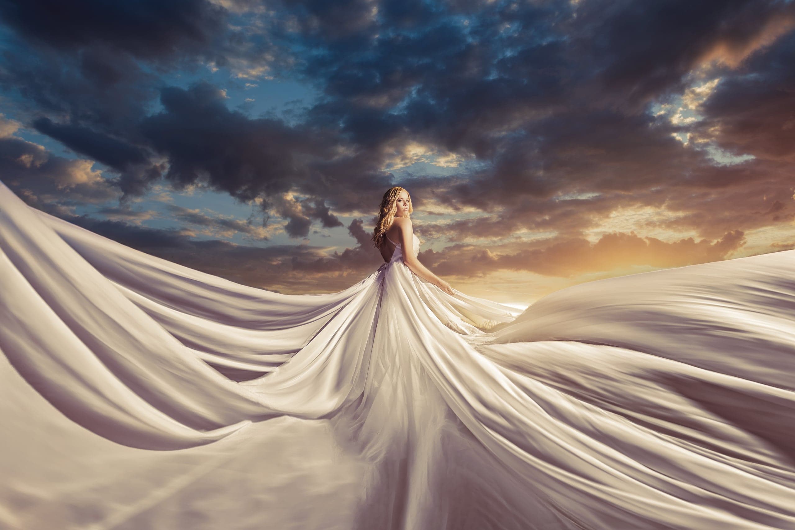 Fine-art senior portrait in a flowing white gown under a dramatic sky