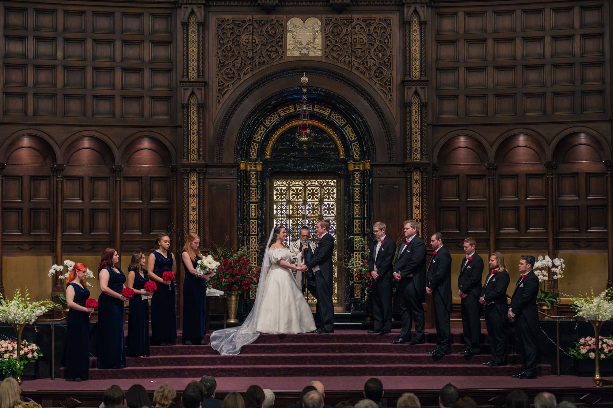 The full wedding party in navy and black formalwear beside the couple at a gilded temple altar