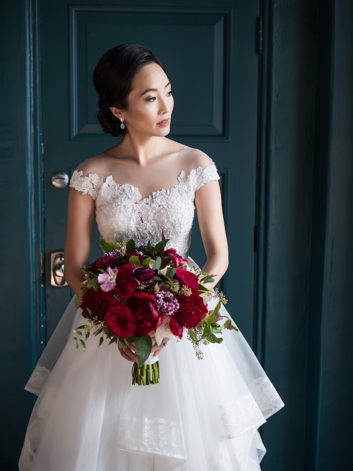Formal bridal portrait against a deep teal door, an off-shoulder lace gown and a burgundy rose bouquet