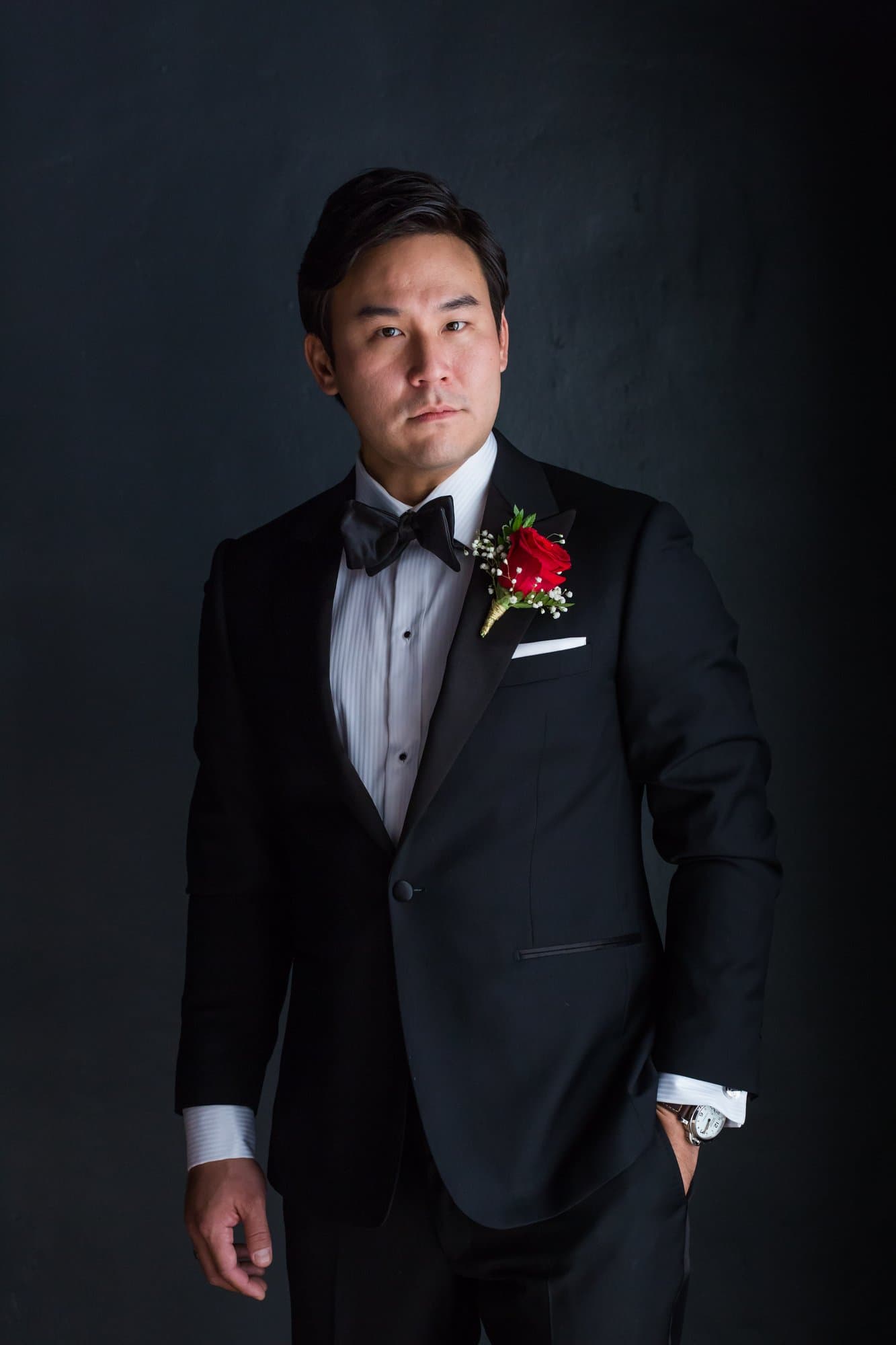 Classic groom portrait in a black tuxedo with a red rose boutonniere against a dark backdrop