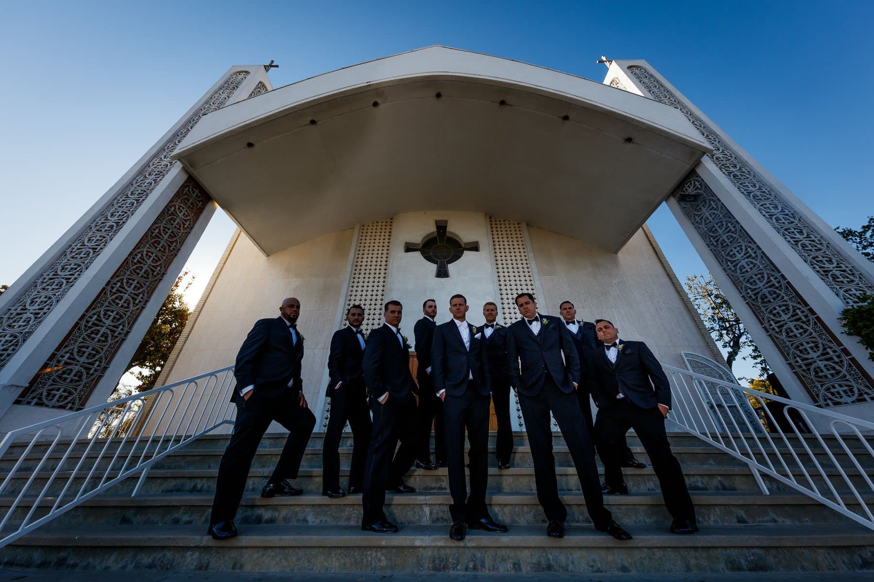 Groomsmen in black tuxedos arranged down the steps of a modern Greek Orthodox church