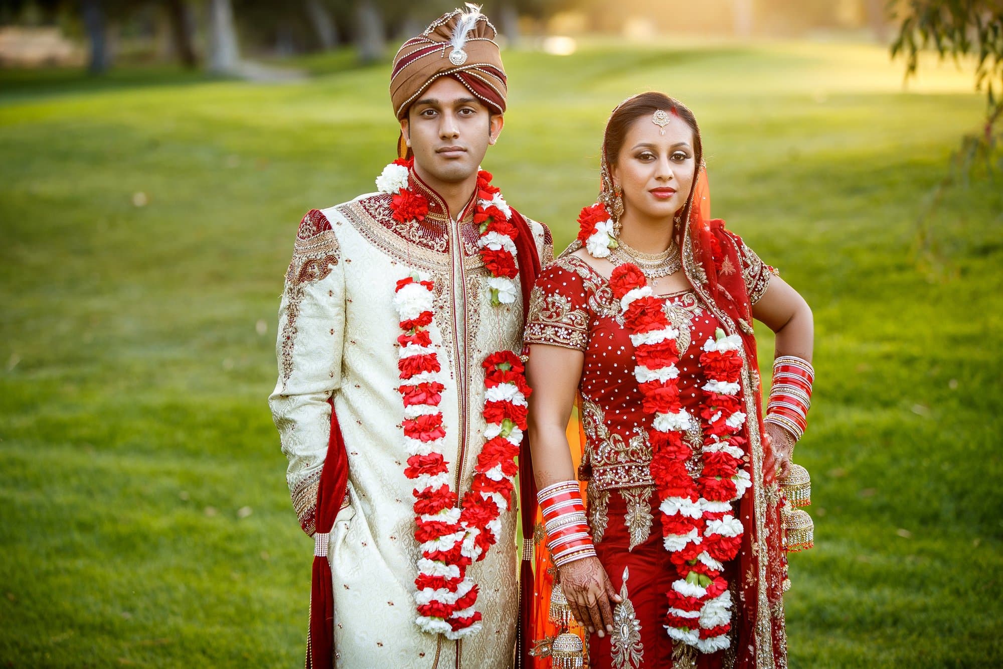 A traditional desi wedding portrait, groom in an ivory sherwani and bride in a red lehenga with floral jaimala garlands at golden hour