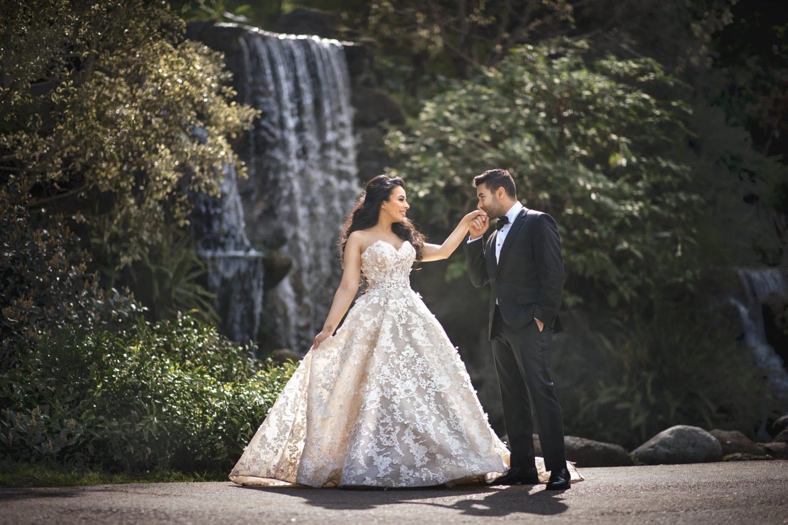 A couple walking a tree-lined path at the Arboretum of Los Angeles County in Arcadia, with lush greenery and a historic garden building behind them.