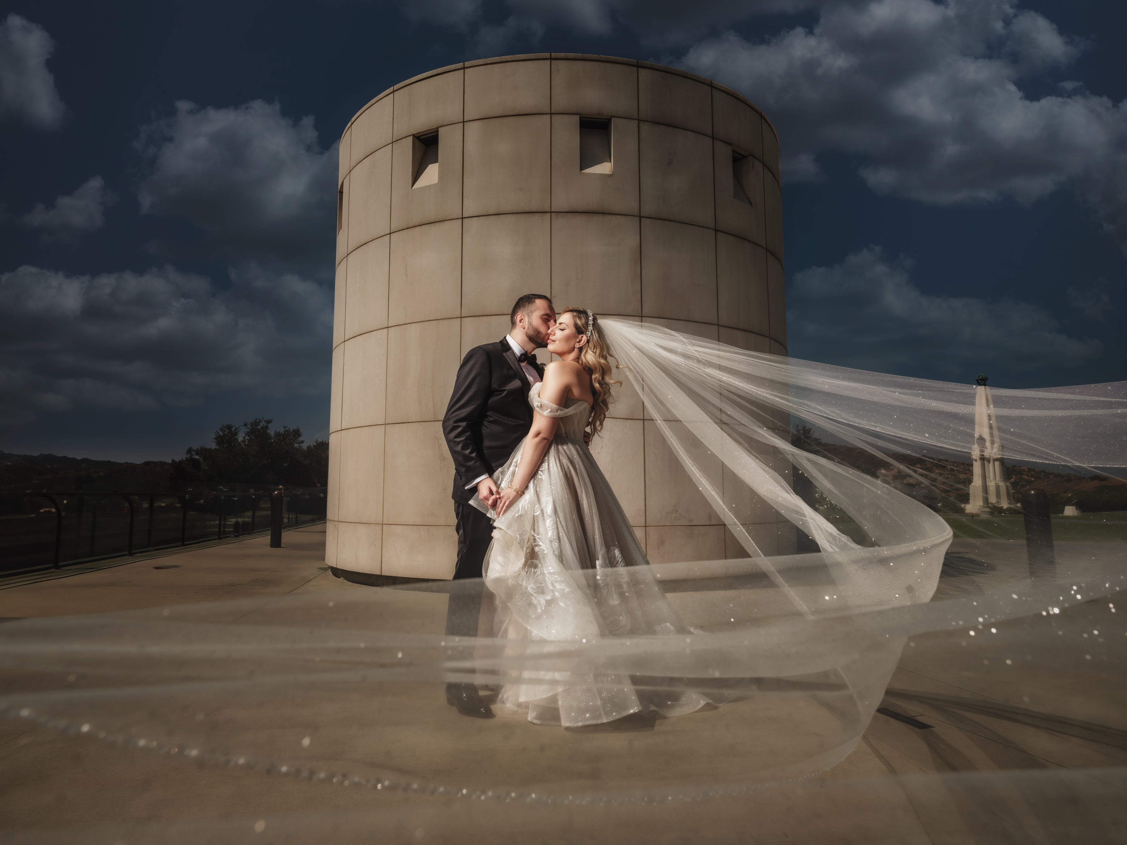 Newlyweds embracing beside modern architecture beneath dramatic clouds