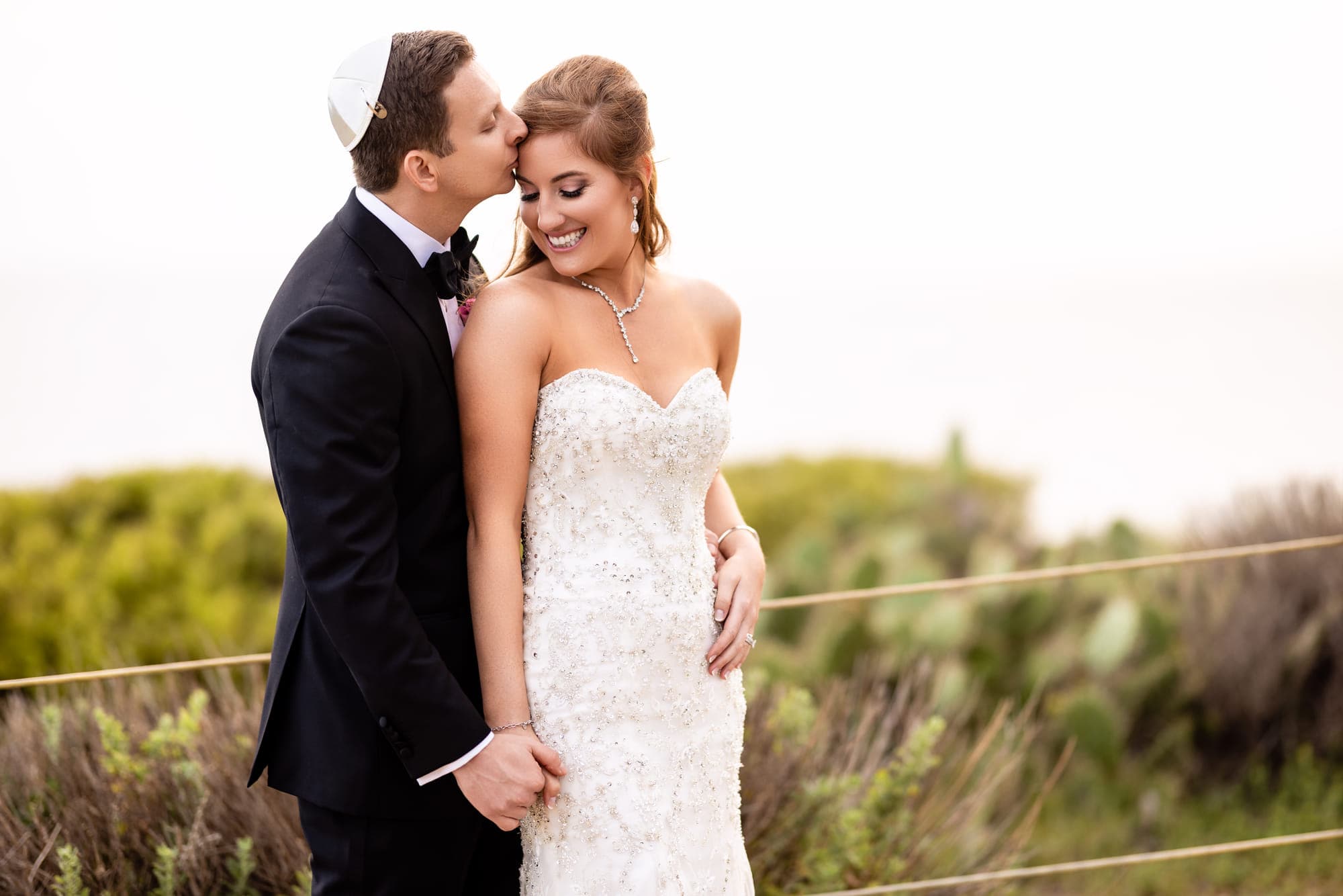 a groom kissing his bride's forehead on a coastal bluff, after retouching