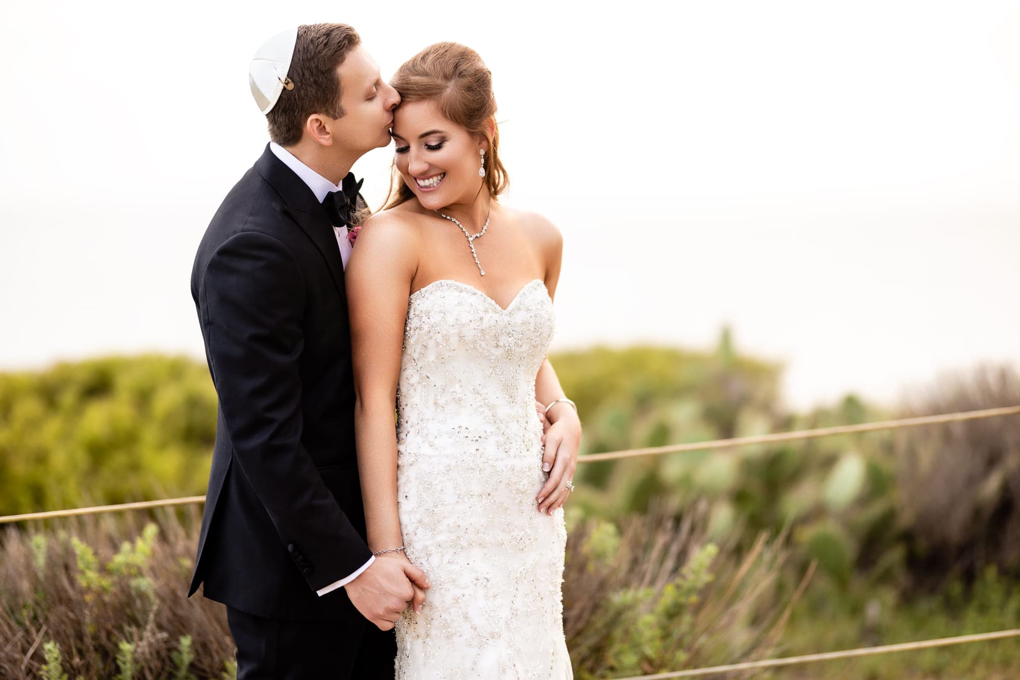 a groom kissing his bride's forehead on a coastal bluff, straight out of camera