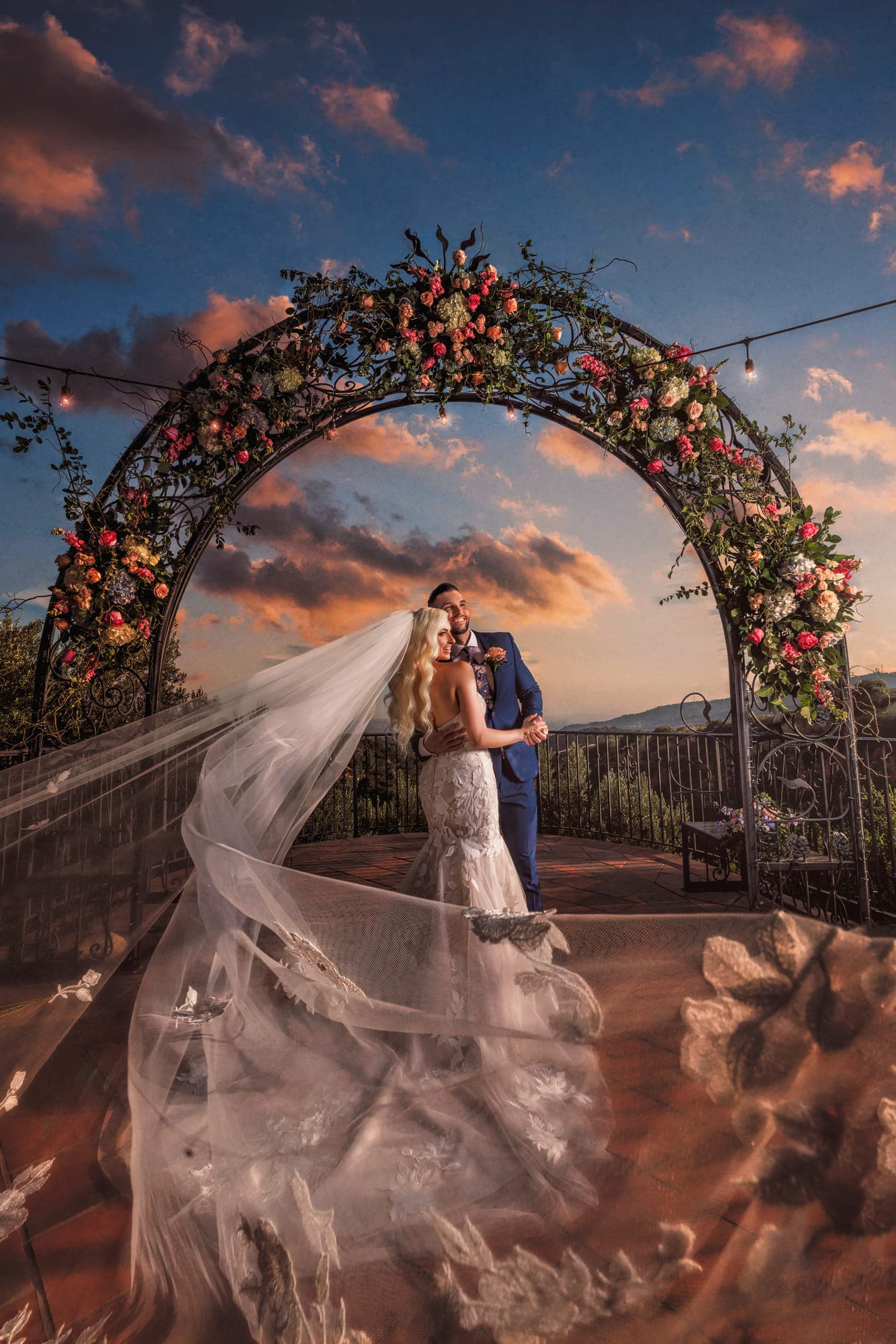 a bride and groom beneath a floral arch at sunset, her veil sweeping across the terrace, after retouching