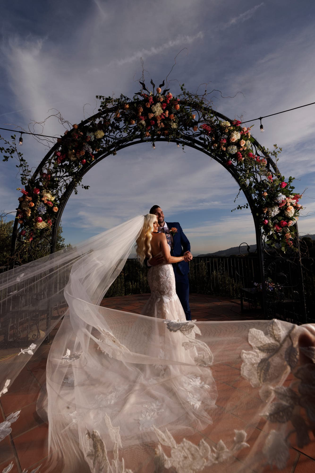 a bride and groom beneath a floral arch at sunset, her veil sweeping across the terrace, straight out of camera