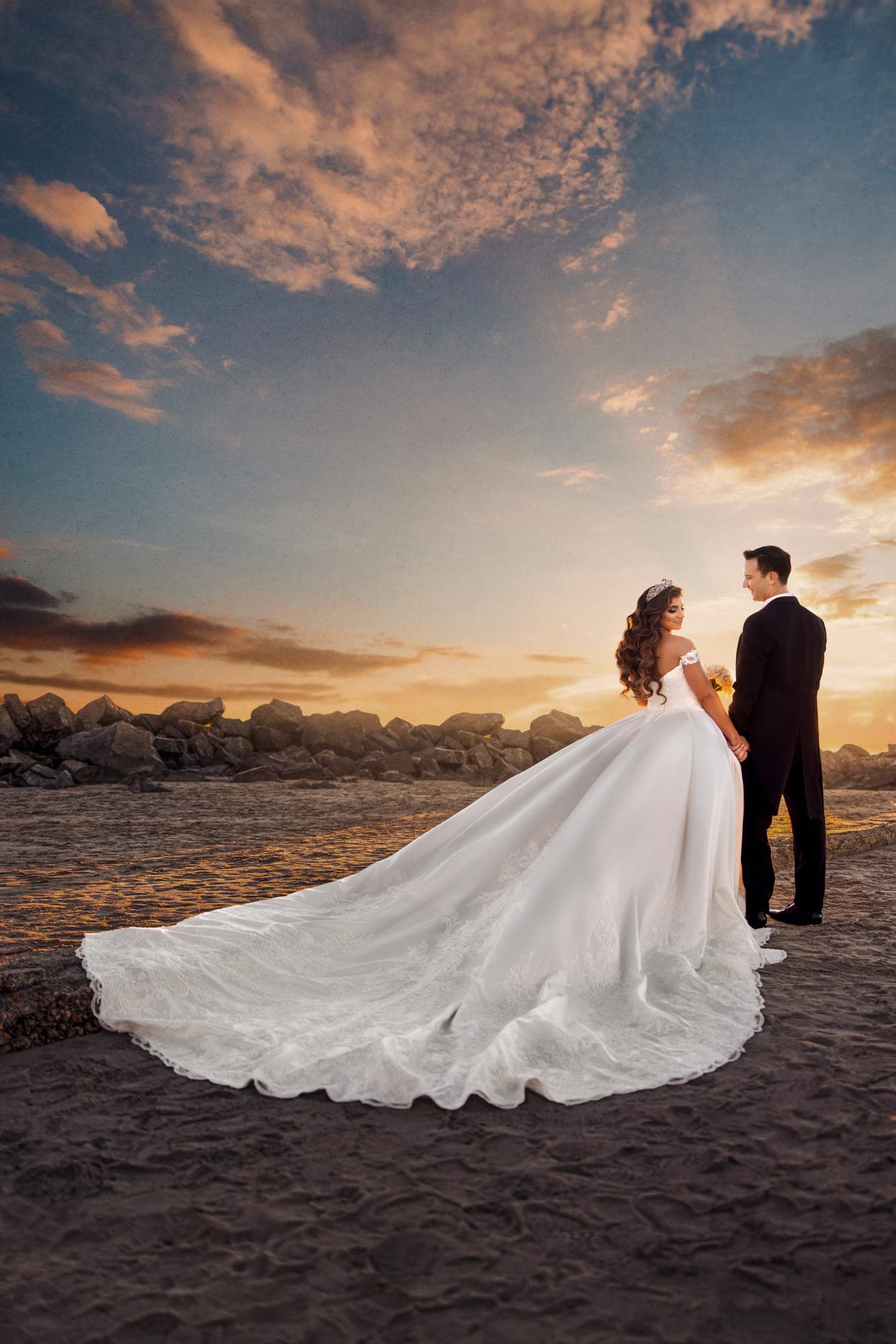 a bride in a full ball gown with the train fanned across the sand, her groom beside her, after retouching