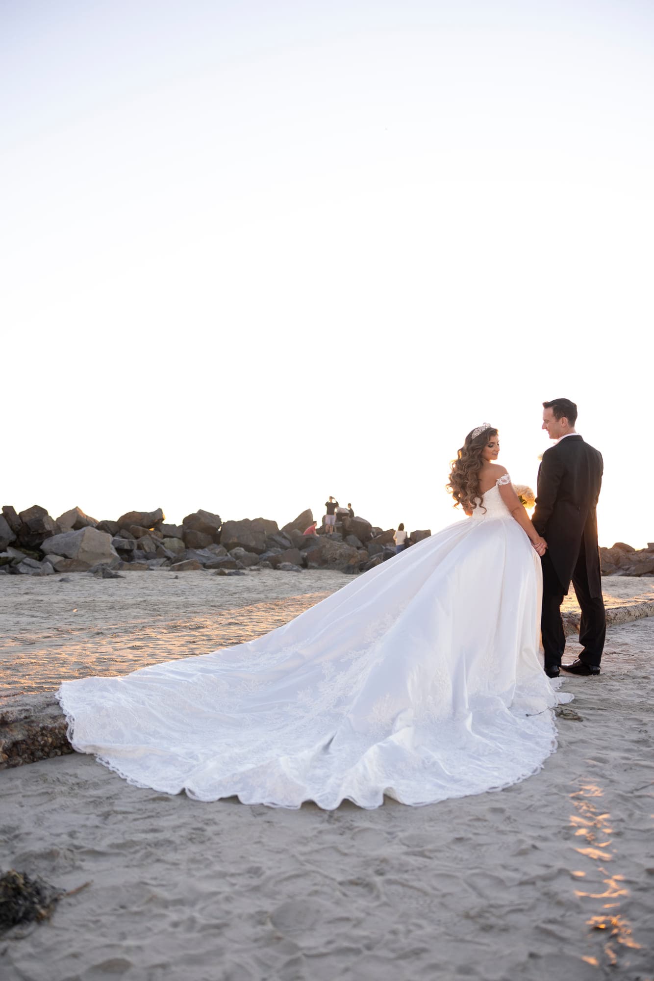 a bride in a full ball gown with the train fanned across the sand, her groom beside her, straight out of camera