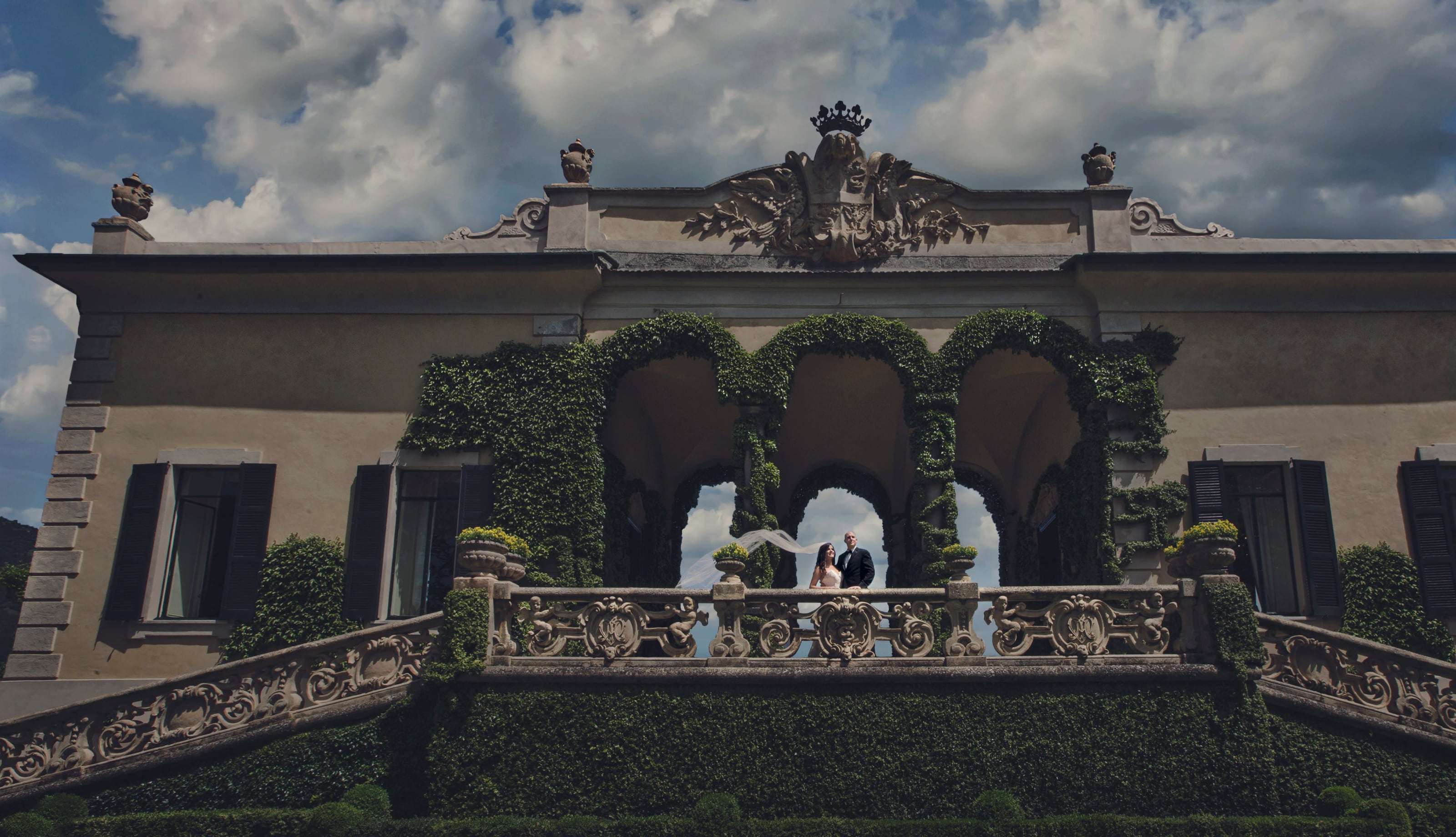 Bride and groom on the baroque balustrade of Villa del Balbianello, Lake Como, her veil catching the wind