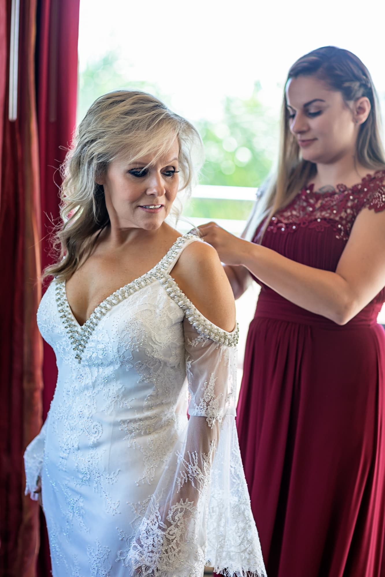 An attendant in burgundy fastening the shoulder of Sheridi's beaded lace gown during prep