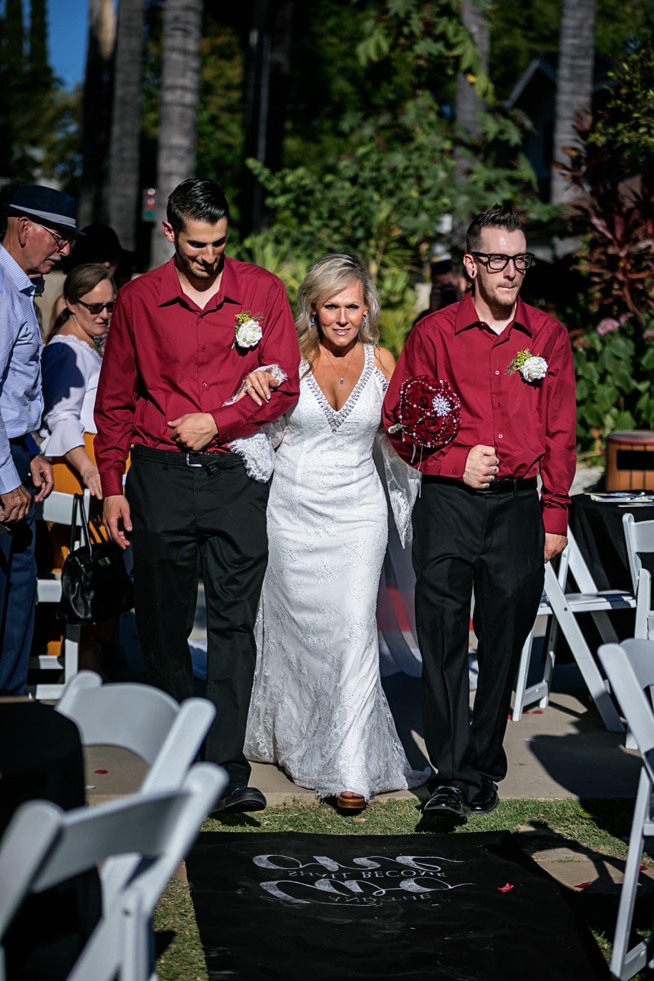 Sheridi walking the garden aisle escorted on both arms by two men in burgundy shirts at 40 Palms Estates