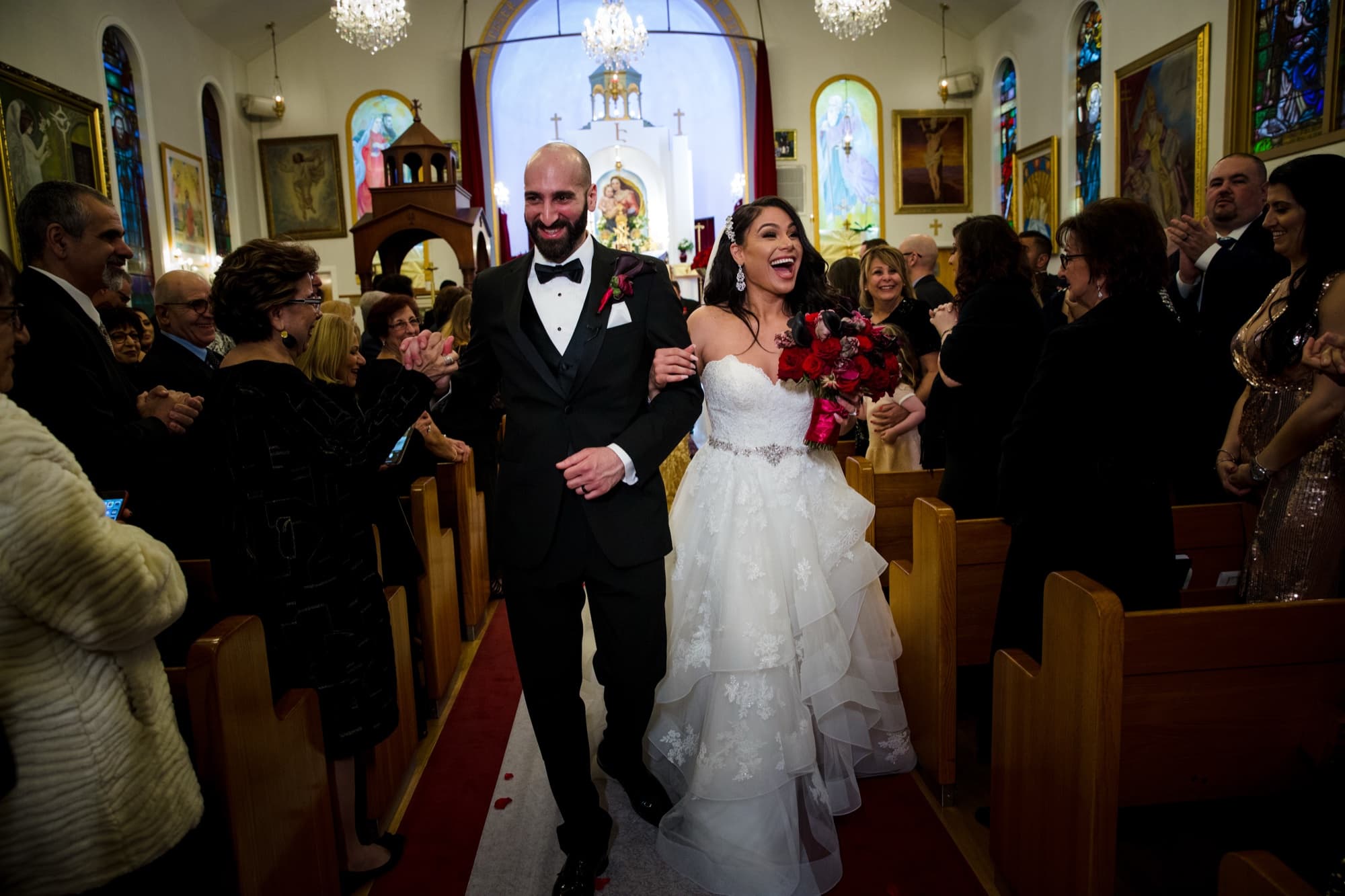 Margaret and Hrug walking back up the aisle of Holy Martyrs Armenian Church past clapping guests, her red rose bouquet raised