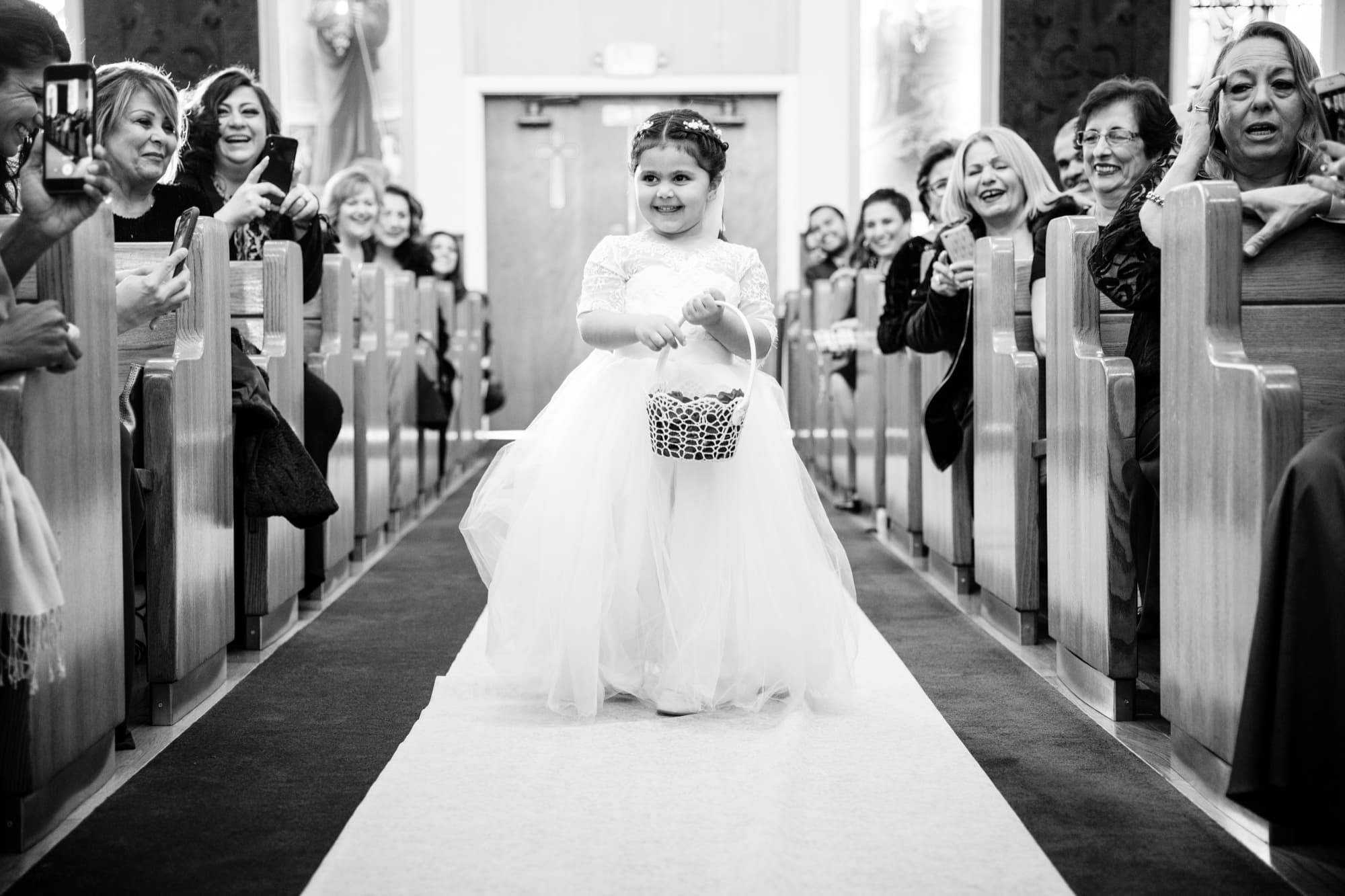 Black and white photograph of the flower girl walking down the church aisle between clapping guests