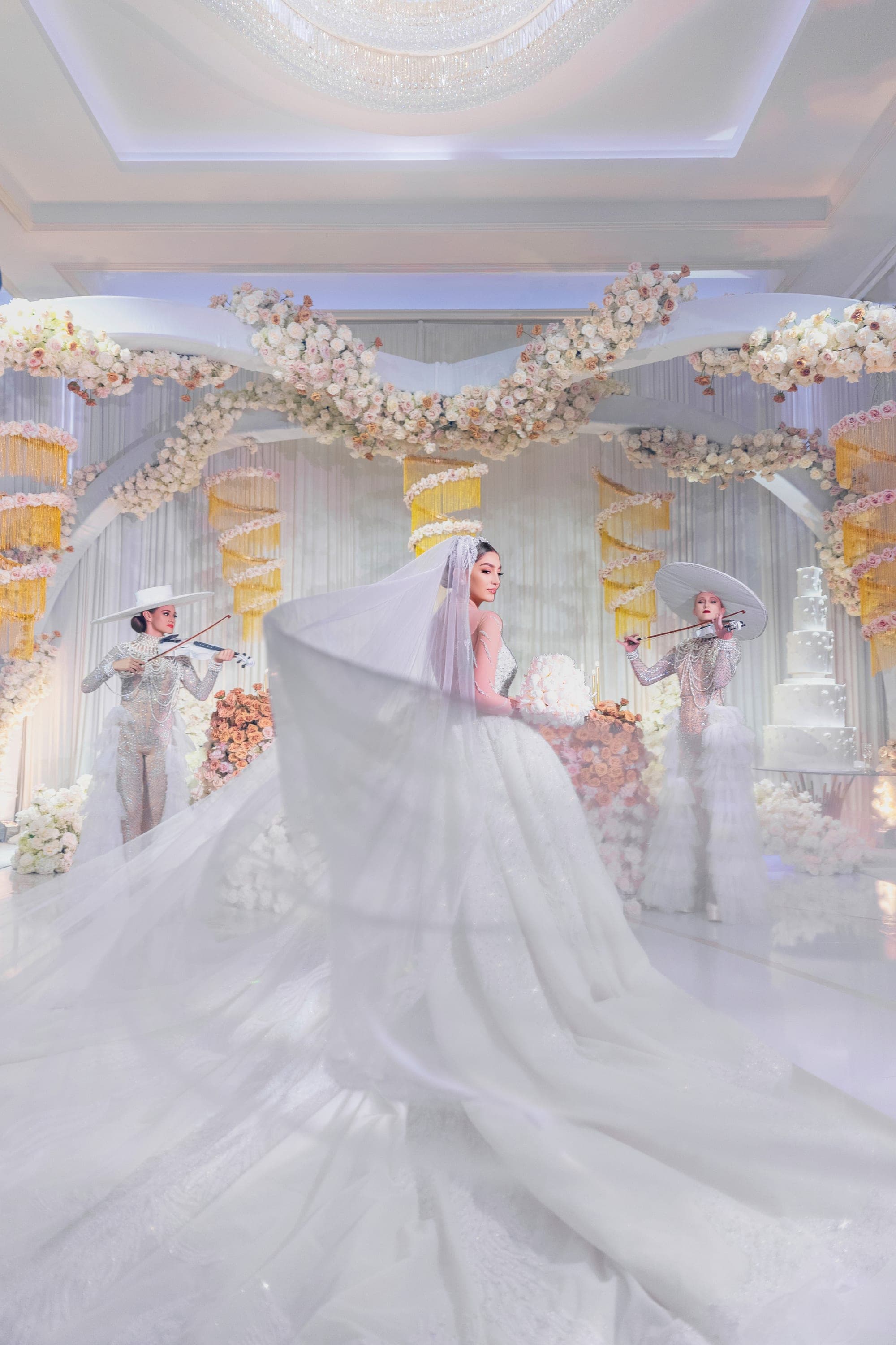 The bride beneath her cathedral veil in the ballroom, flanked by violinists in white wide-brimmed hats
