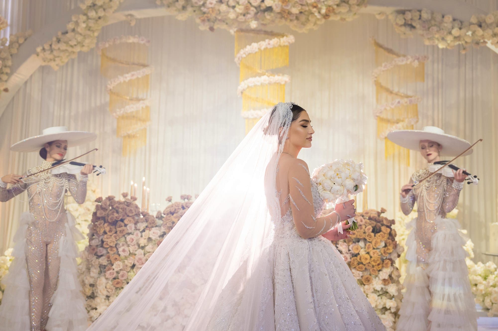 The bride crossing the ballroom floor between two violinists in white wide-brimmed hats