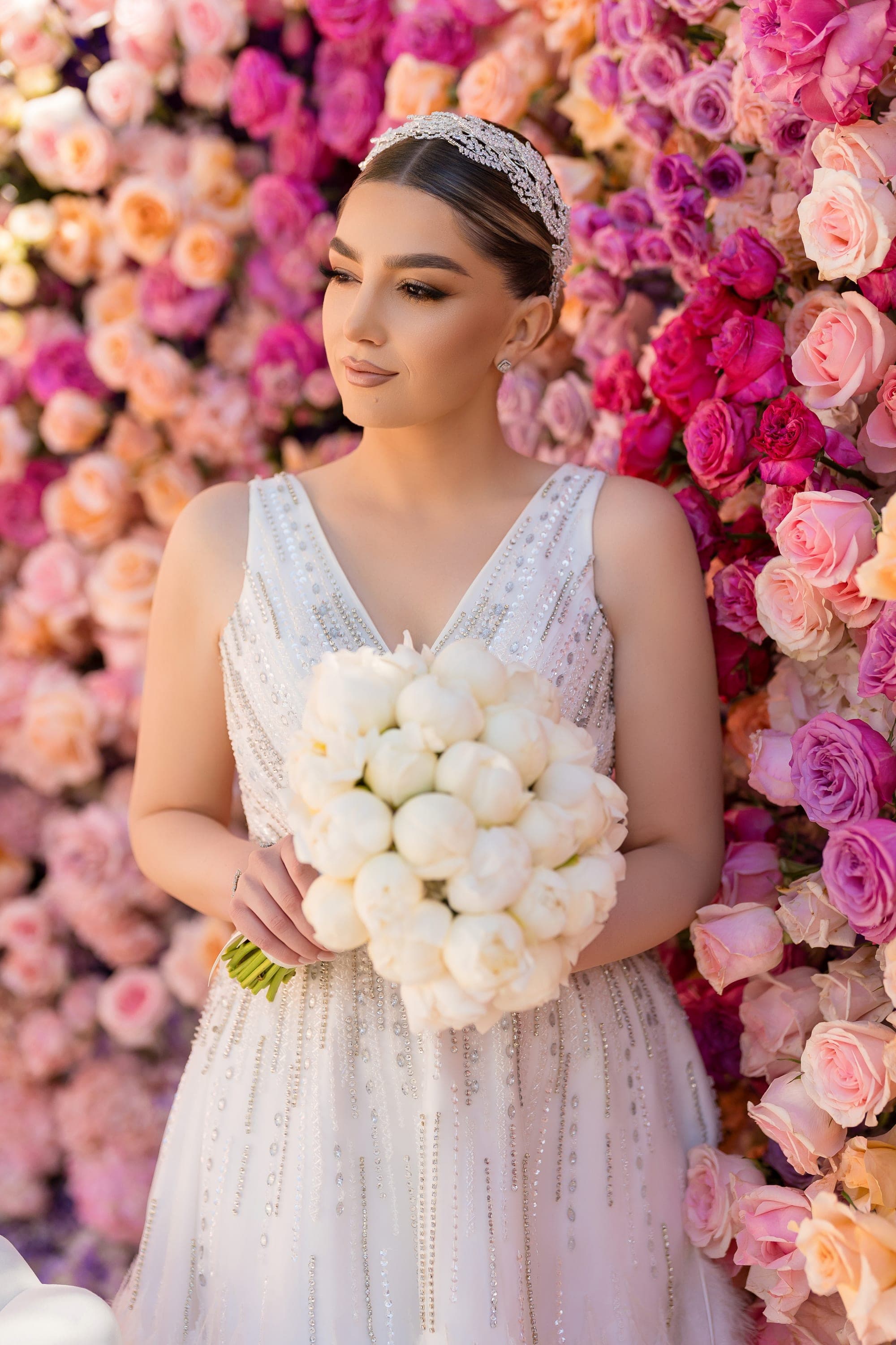 The bride holding a white bouquet in front of a floor-to-ceiling wall of pink and coral roses