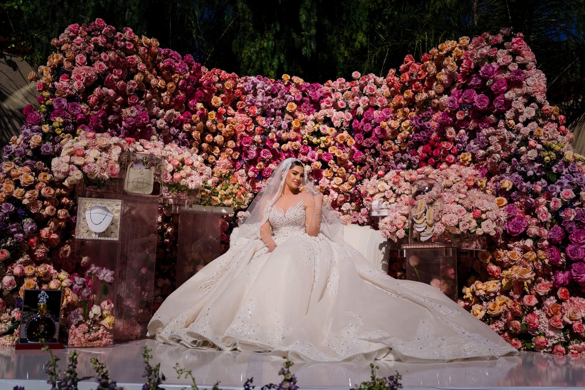 The bride seated in her beaded ballgown before the ombré rose wall with the wedding gifts displayed alongside her