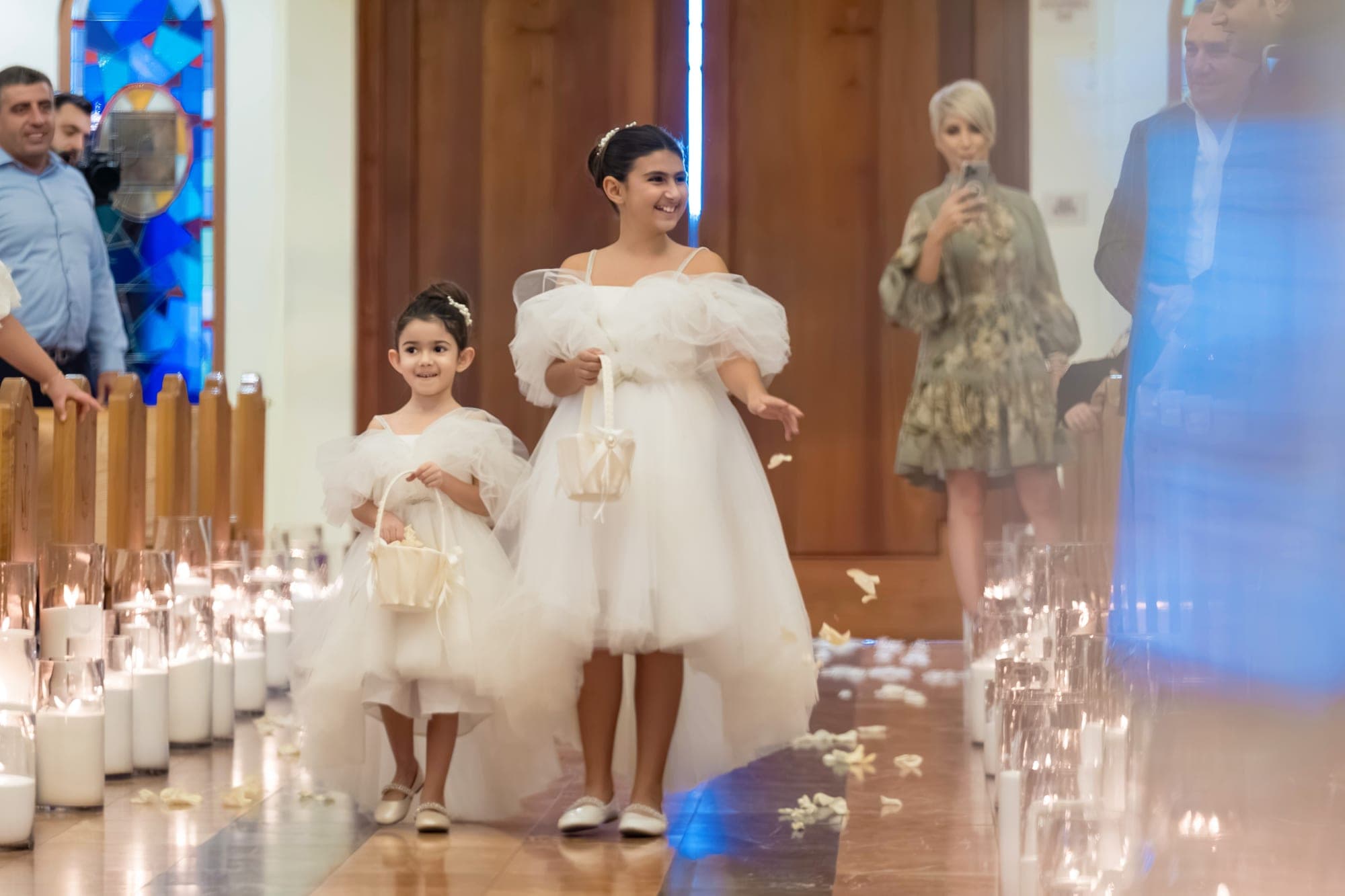 Flower girls in white walking a candlelit church aisle before the ceremony