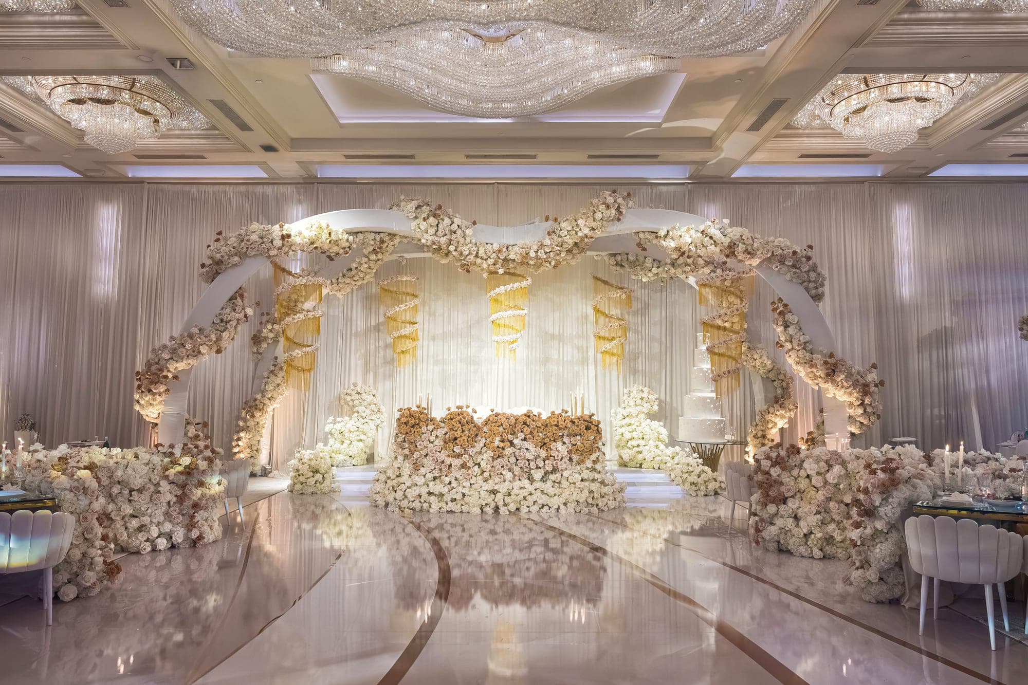 A wedding ballroom in ivory and gold, floral arches and gold ribbon above a mirrored floor