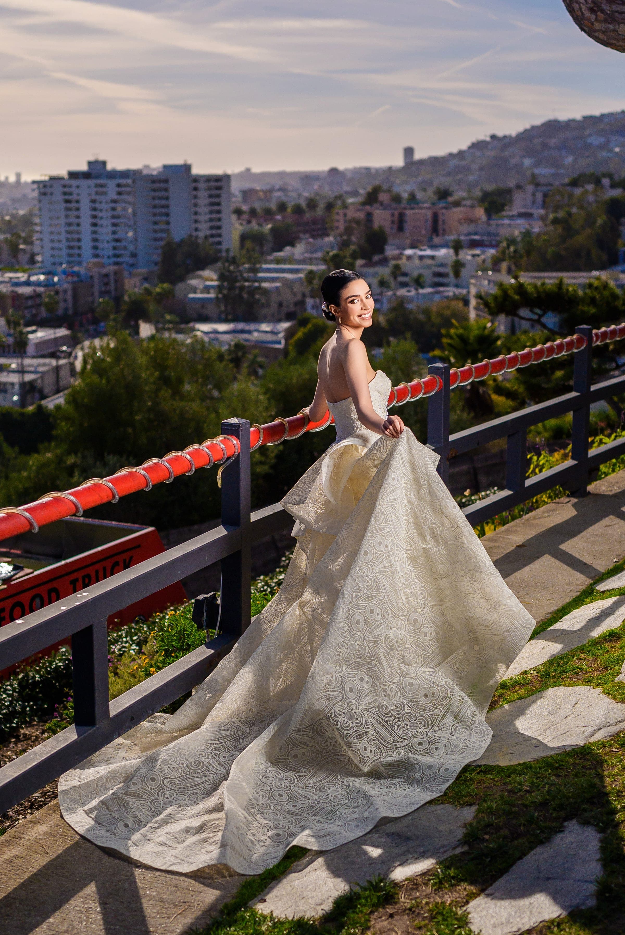 The bride glances back over her shoulder, her lace train spilling down a stone path above the city.
