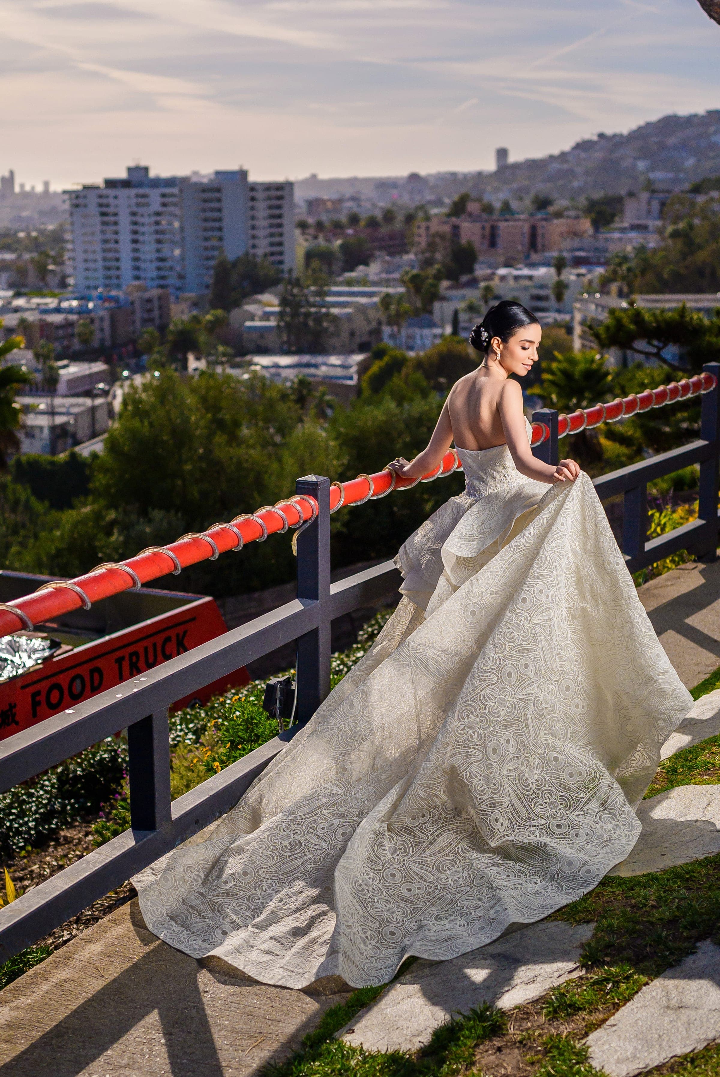 The bride rests a hand on a red railing, her patterned gown fanned out over the hillside overlook.