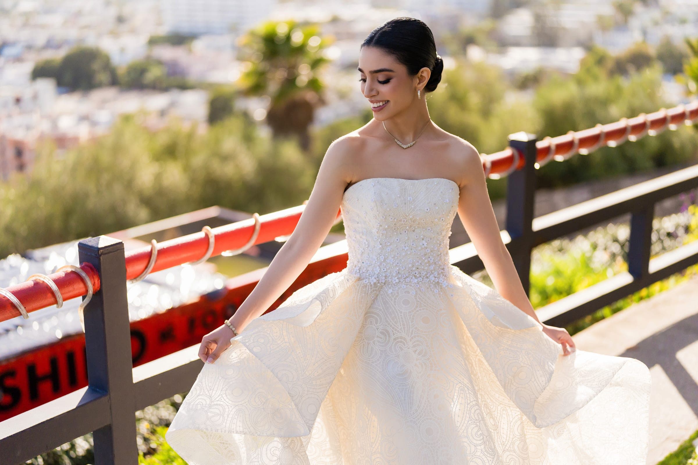 Smiling with eyes lowered, the bride lifts the layered skirt of her gown beside a red railing.