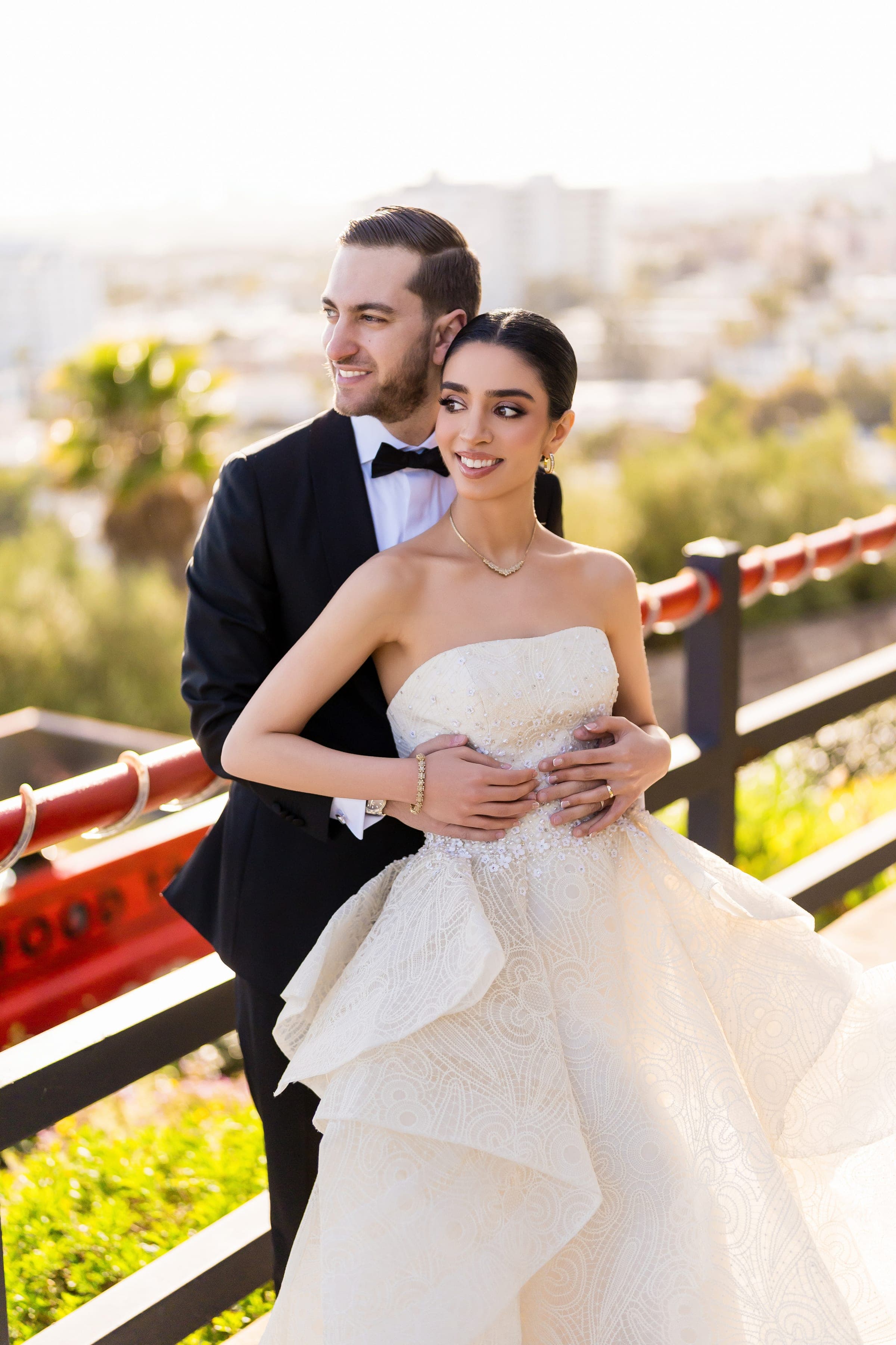 The groom wraps his arms around the bride from behind as they gaze toward the sunlit city.