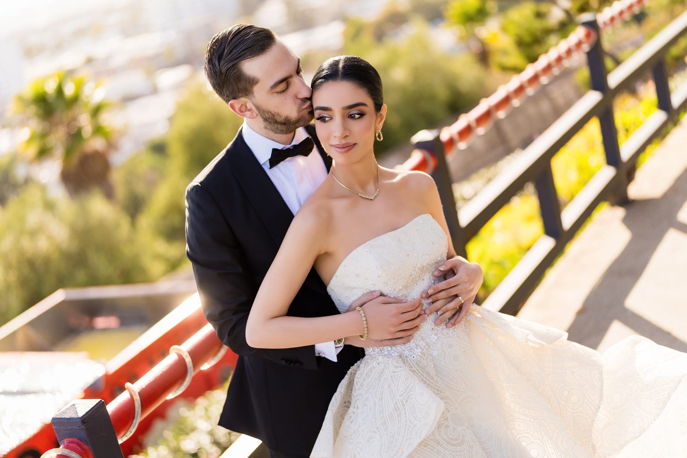 The groom kisses the bride's temple as she smiles softly at the hilltop overlook railing.