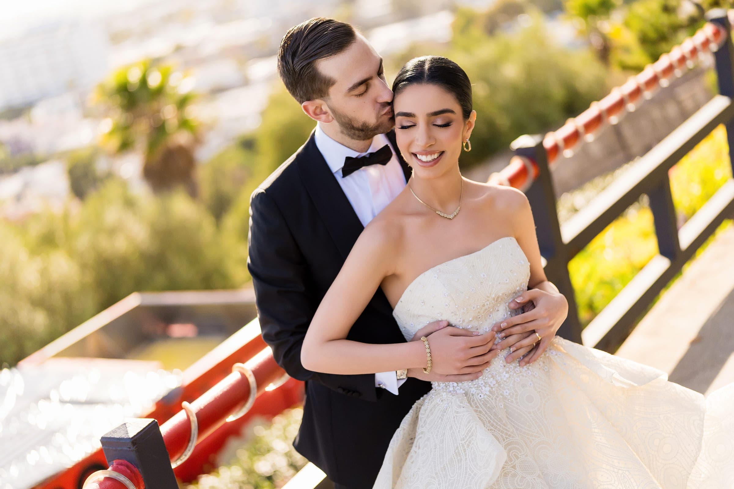 Eyes closed and beaming, the bride laughs as the groom kisses her temple in warm afternoon light.