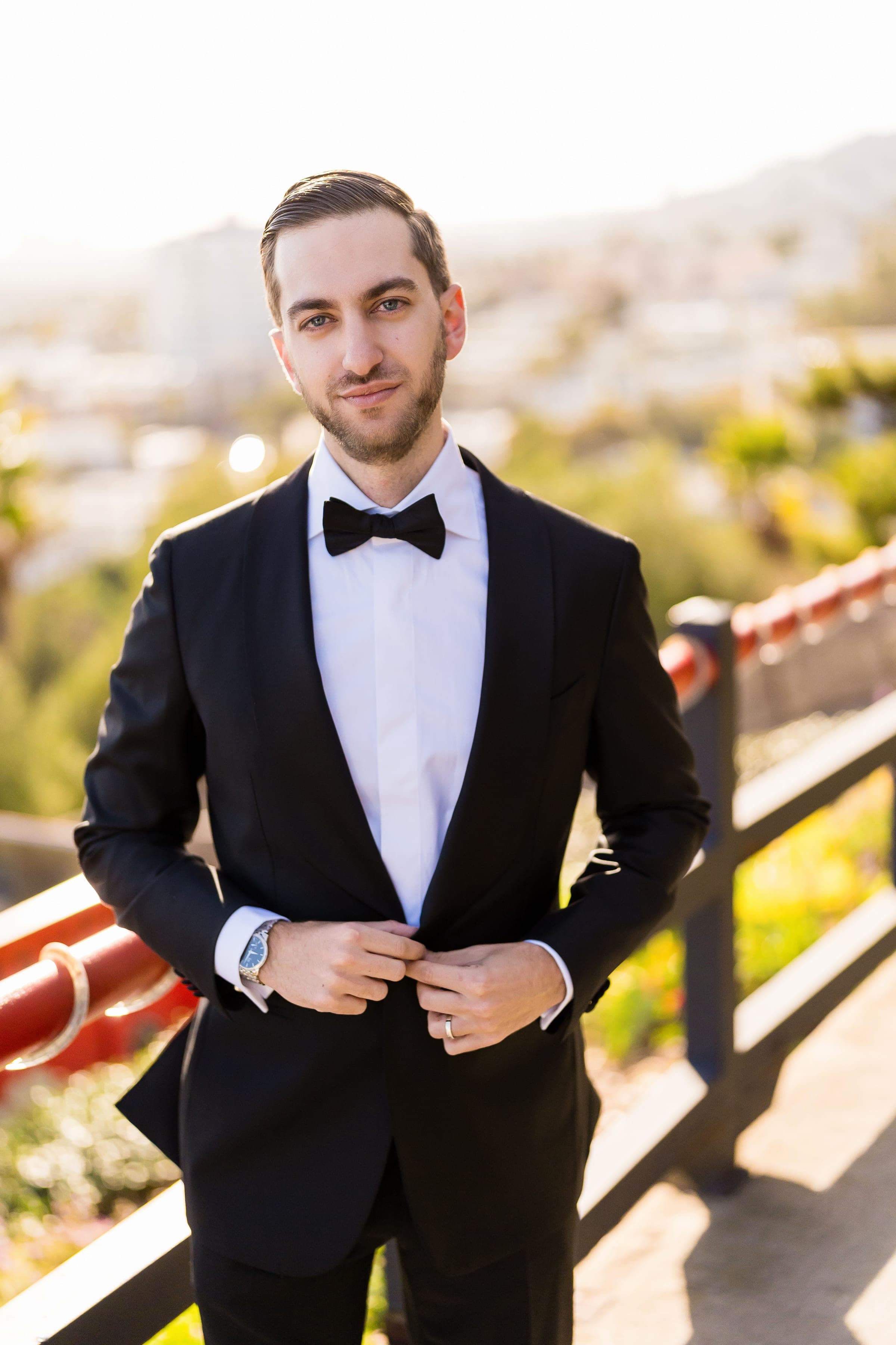 The groom fastens his tuxedo button on a sunlit terrace above the hazy city below.