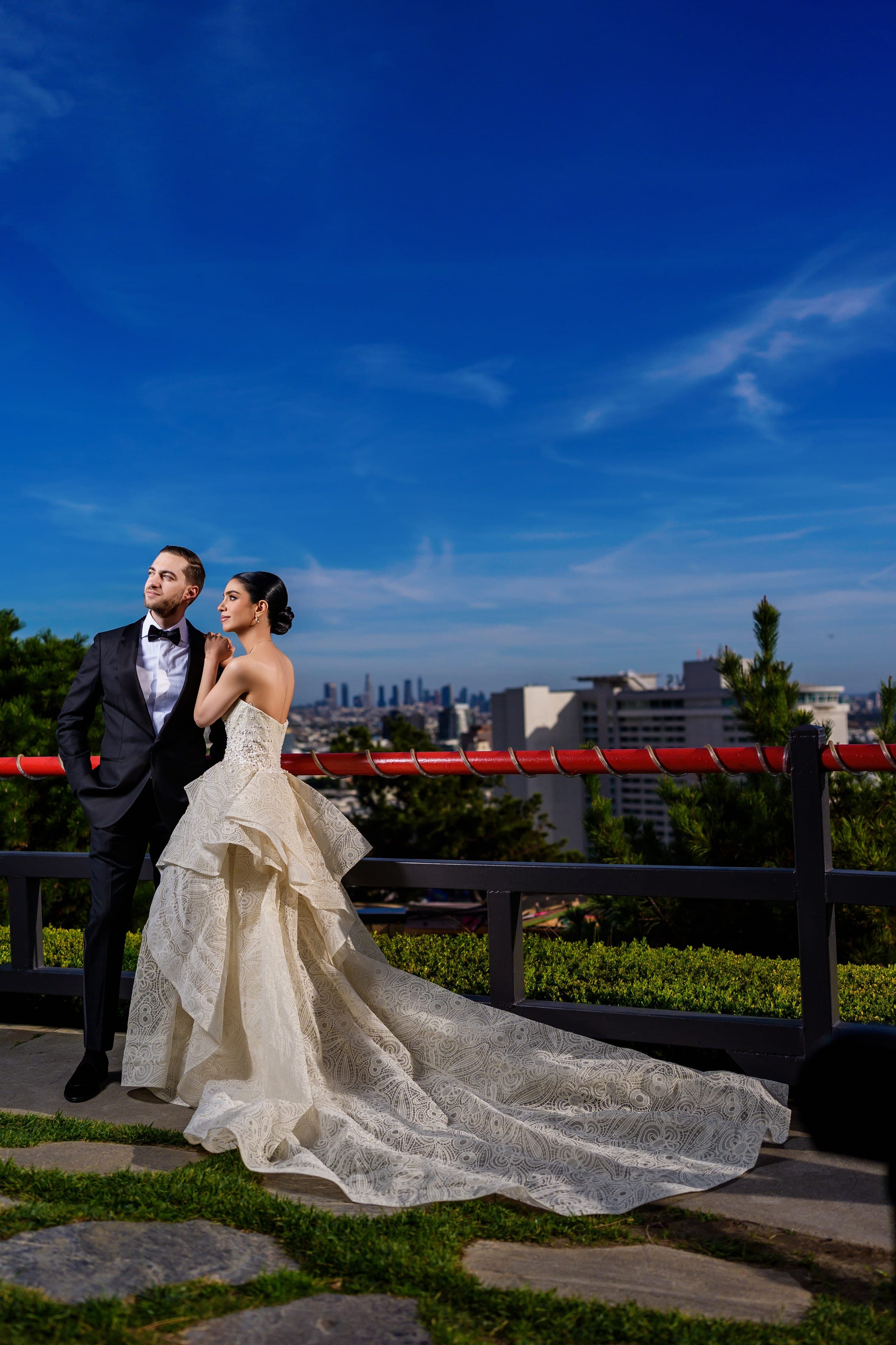 The bride leans on the groom's shoulder at a red-railed overlook facing the Los Angeles skyline.
