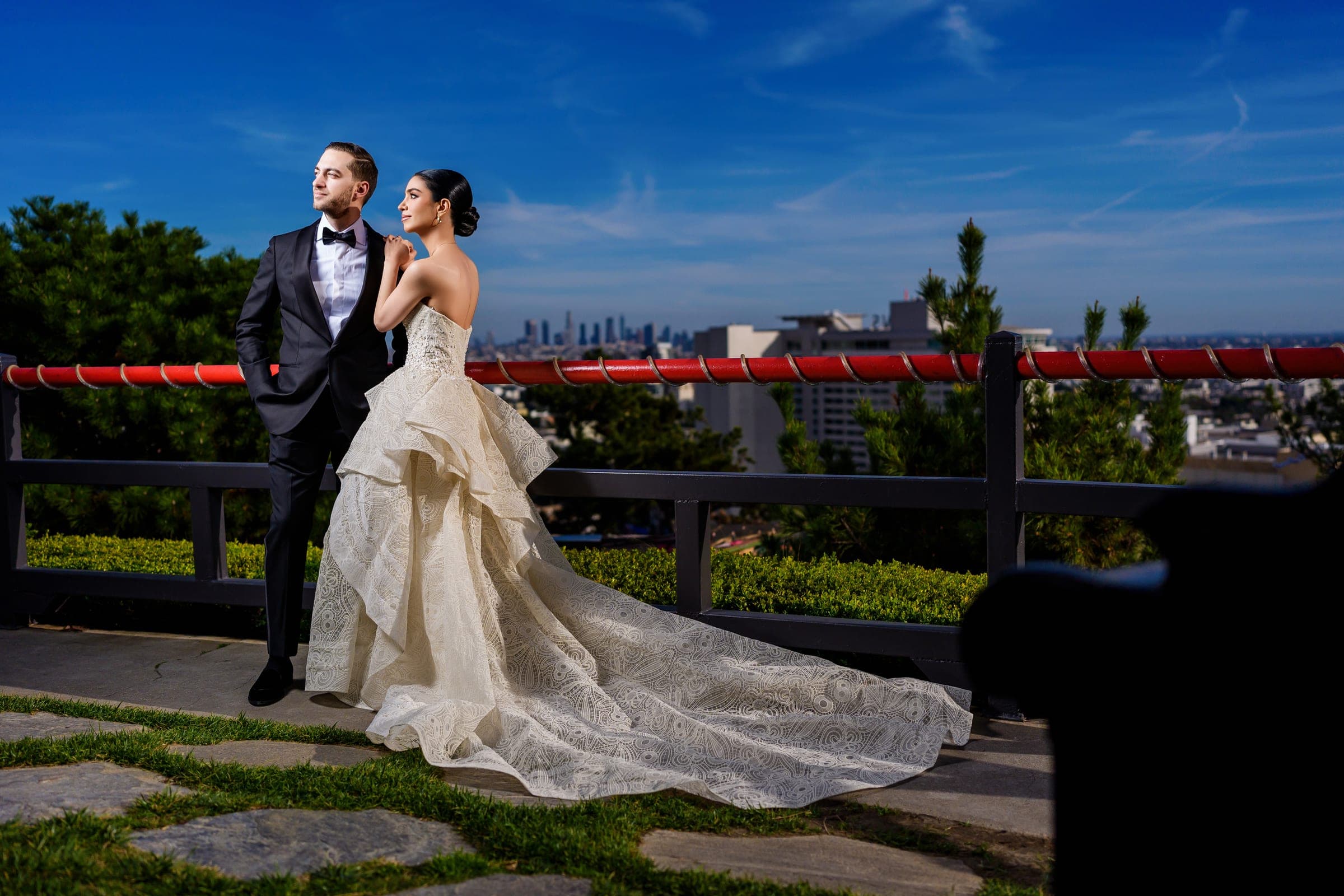 The couple gazes past a red railing toward downtown, her lace train covering the stone path.