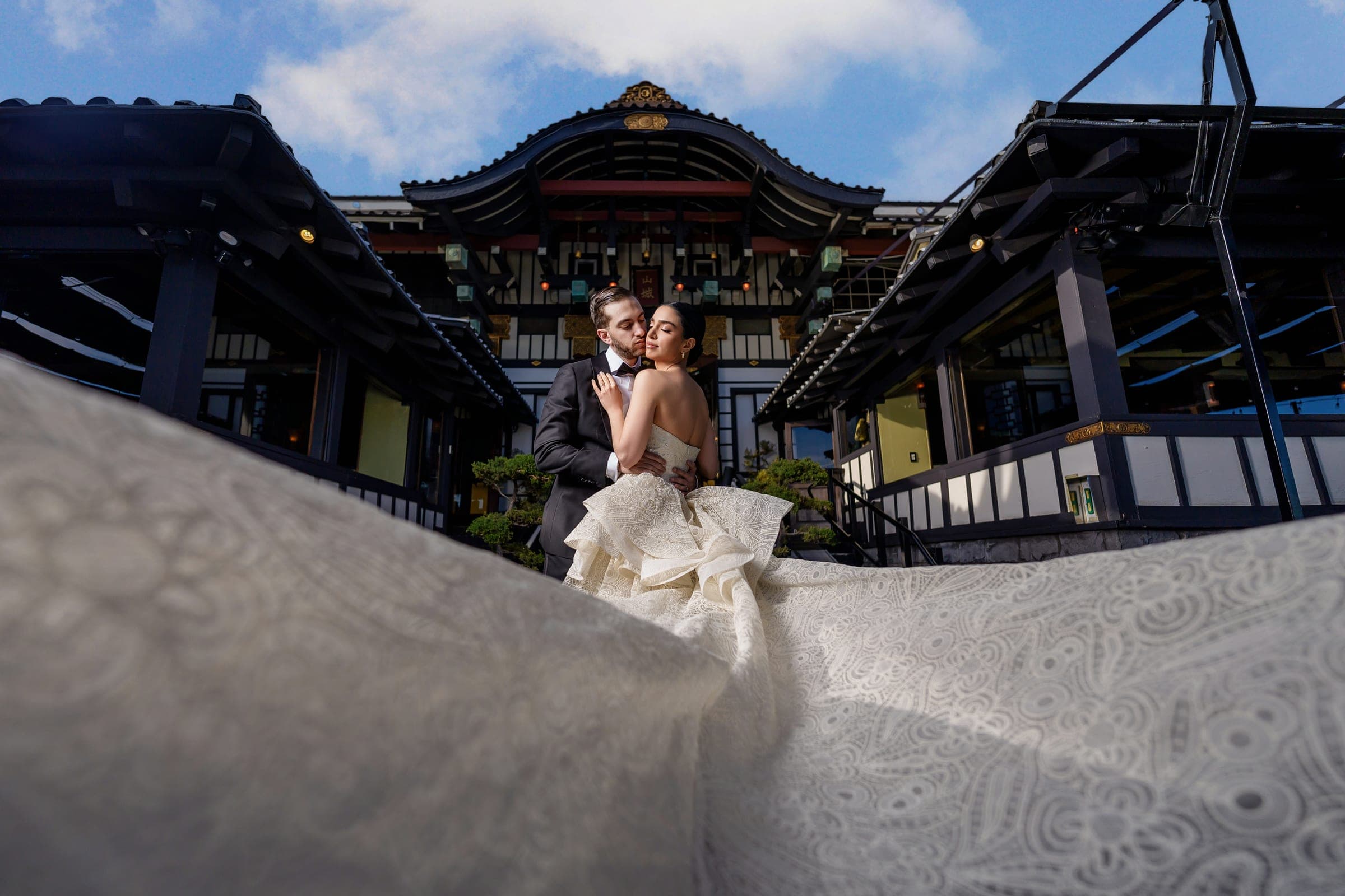 The groom kisses the bride's cheek outside a pagoda-style pavilion, her lace train sweeping into the foreground.