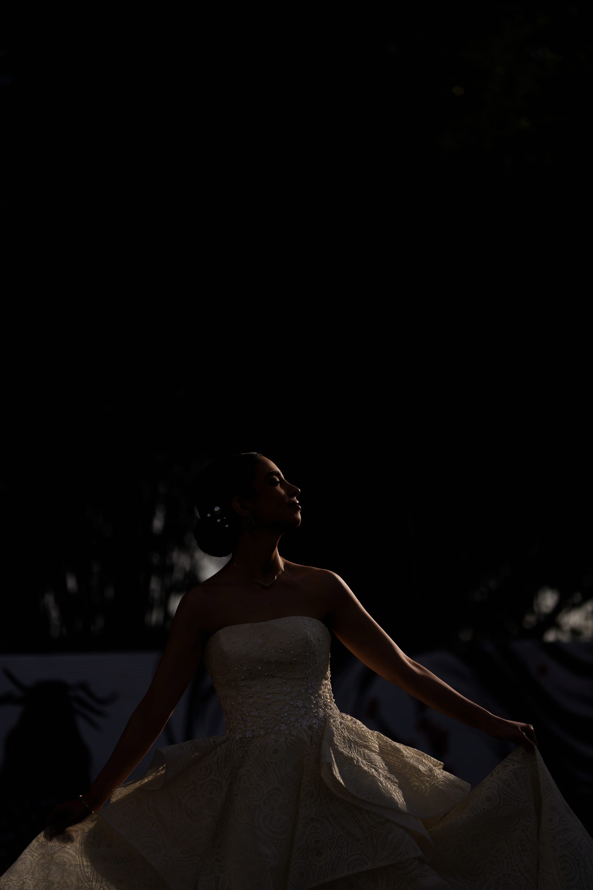 Rim light outlines the bride's profile and bare shoulders as she fans her skirt in darkness.