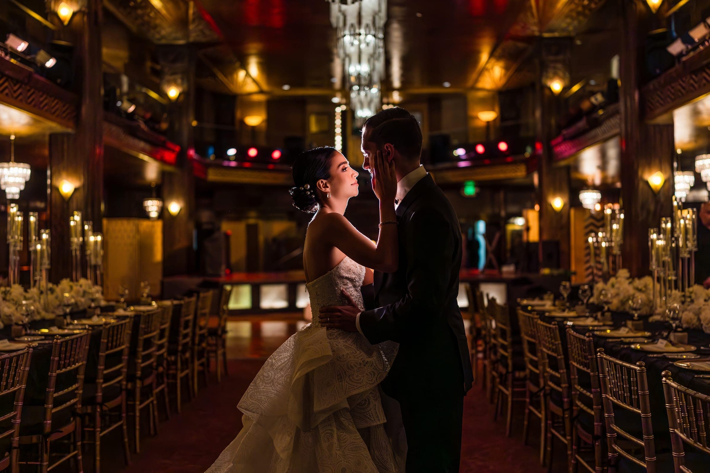 The bride touches the groom's cheek between candlelit tables in a gilded Art Deco ballroom.