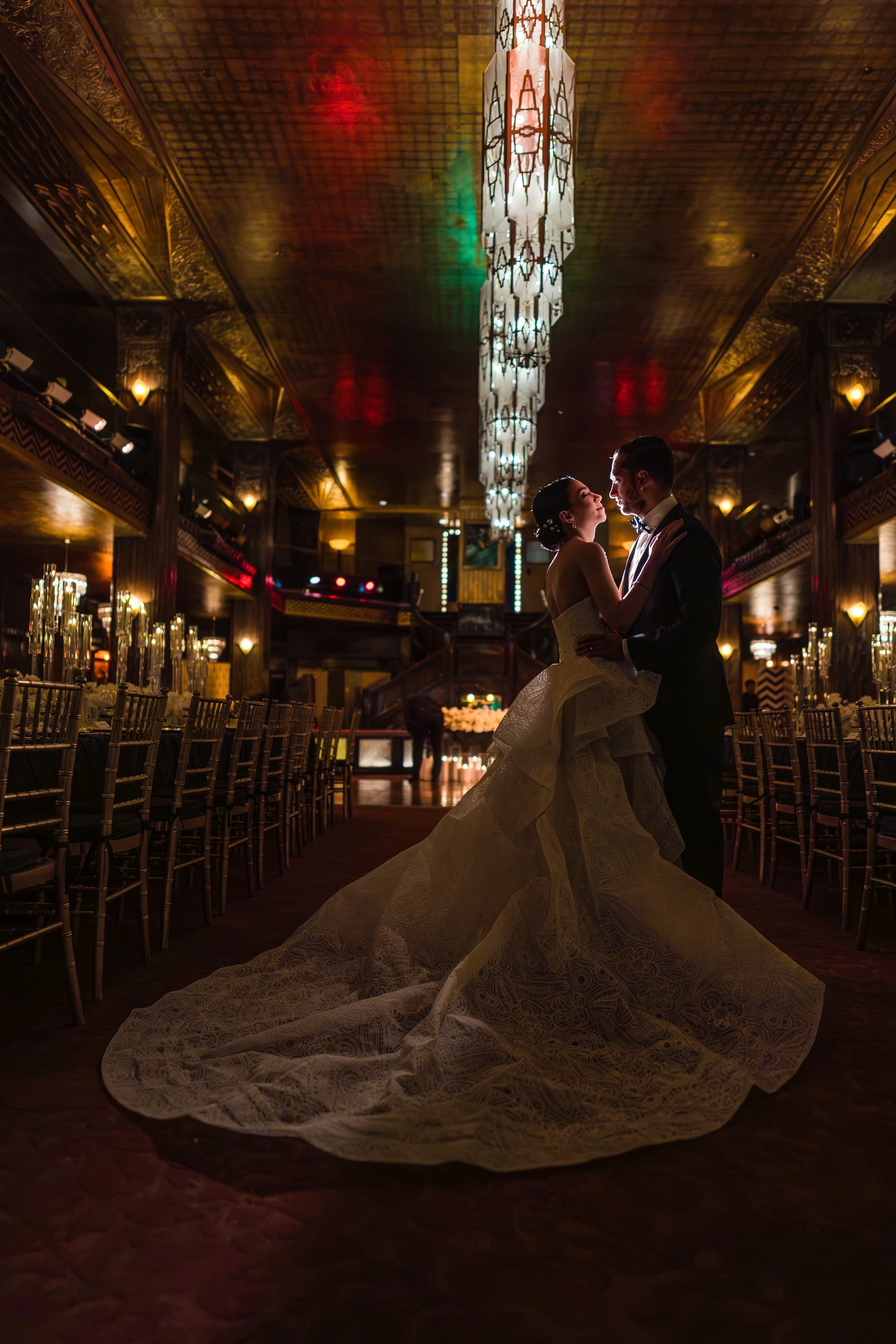 The couple embraces beneath a tiered Art Deco chandelier in Cicada's gold-ceilinged ballroom, her lace train pooling behind.