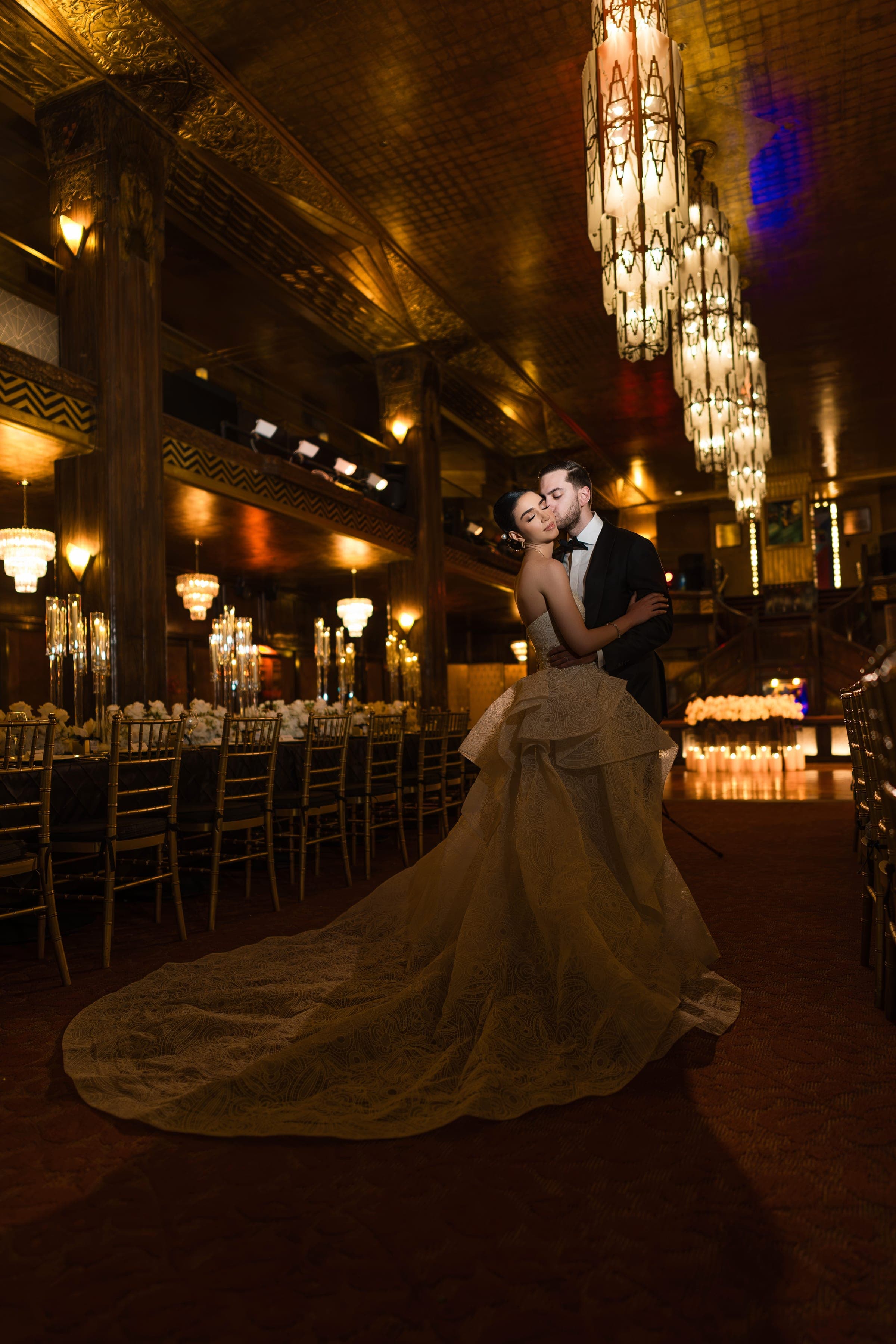 The groom kisses the bride's cheek beside long candlelit banquet tables under Cicada's glowing cascading chandeliers.