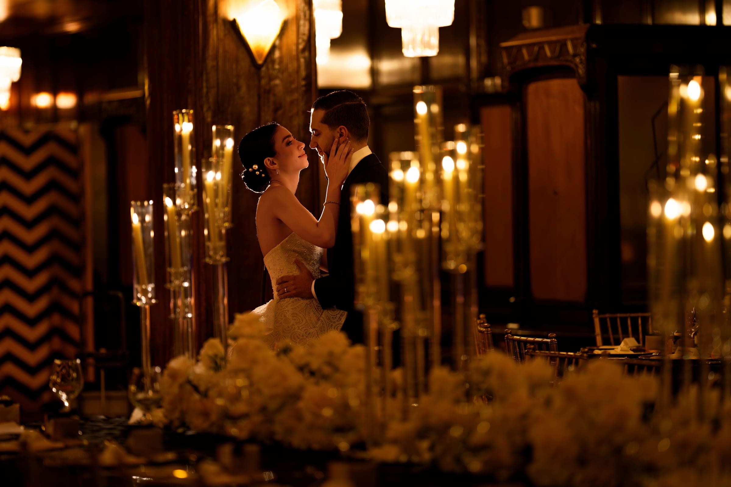 The bride cradles the groom's face amid tall taper candles glowing across the reception tables.