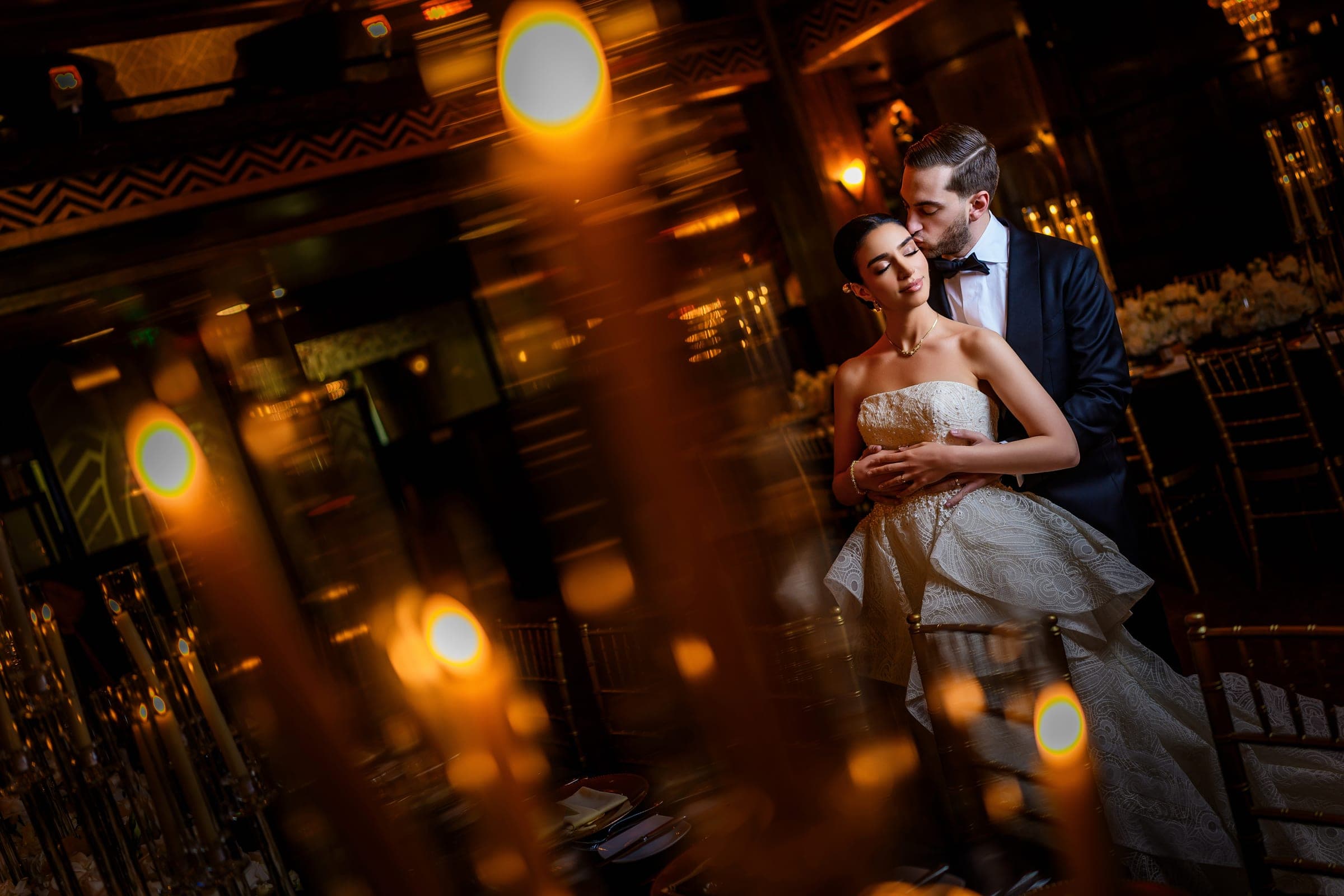Seen through blurred candle flames, the groom kisses the bride's temple in the wood-paneled dining room.