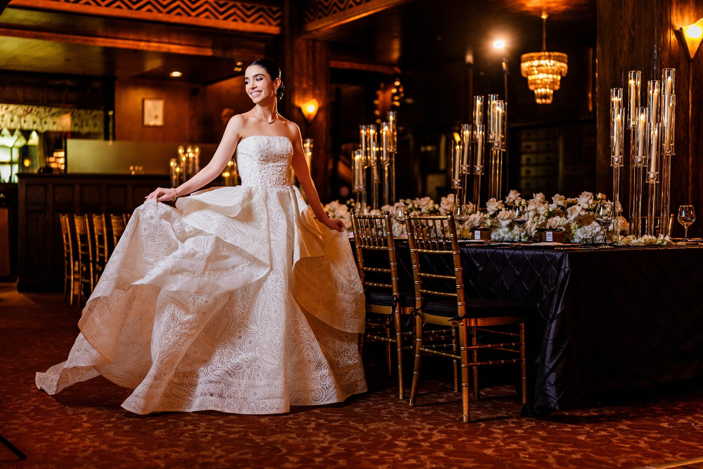 The bride smiles and fans out her patterned ball gown beside a rose-lined reception table and gold chairs.