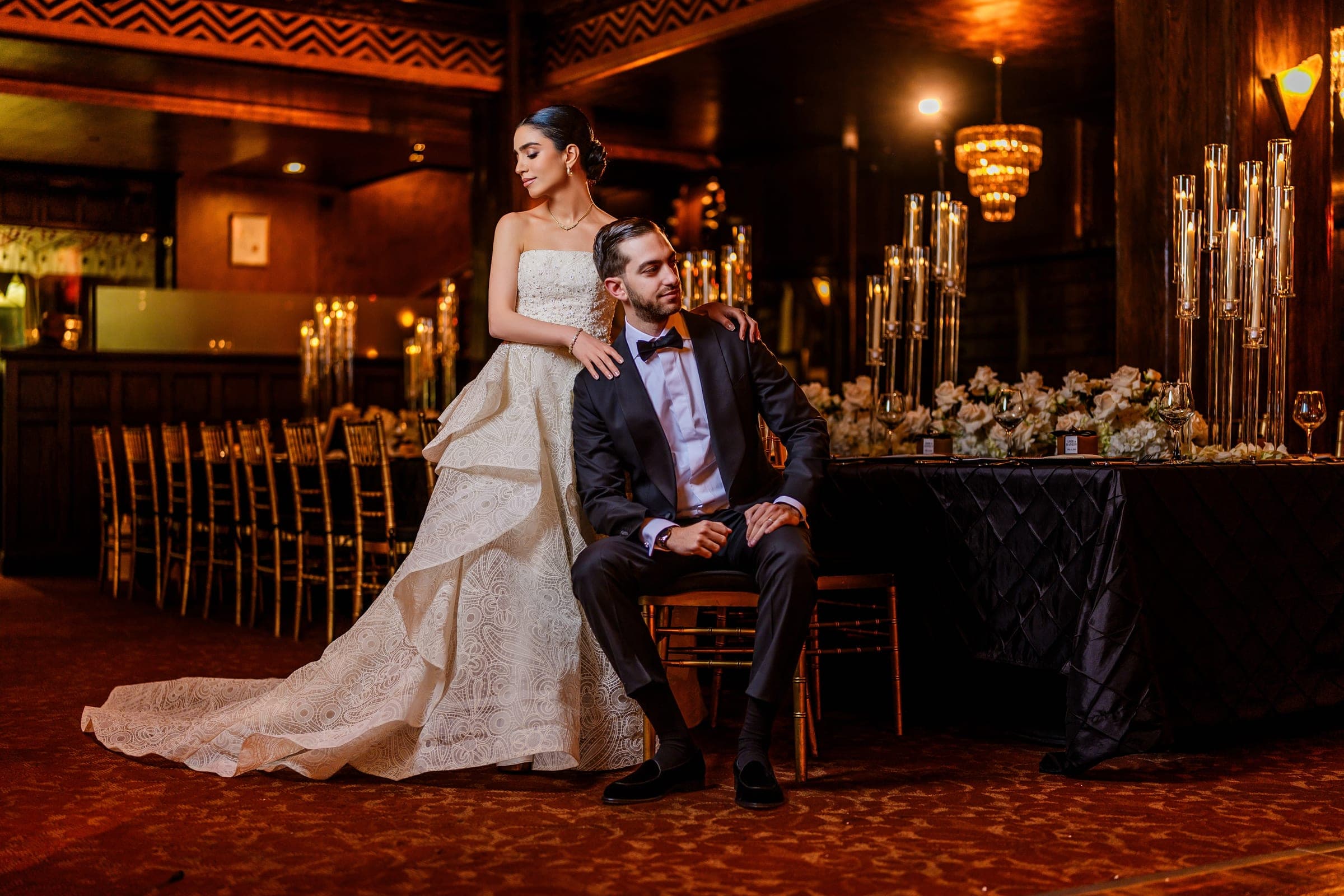 The bride rests her hands on the seated groom's shoulders, surrounded by candelabras and white roses.