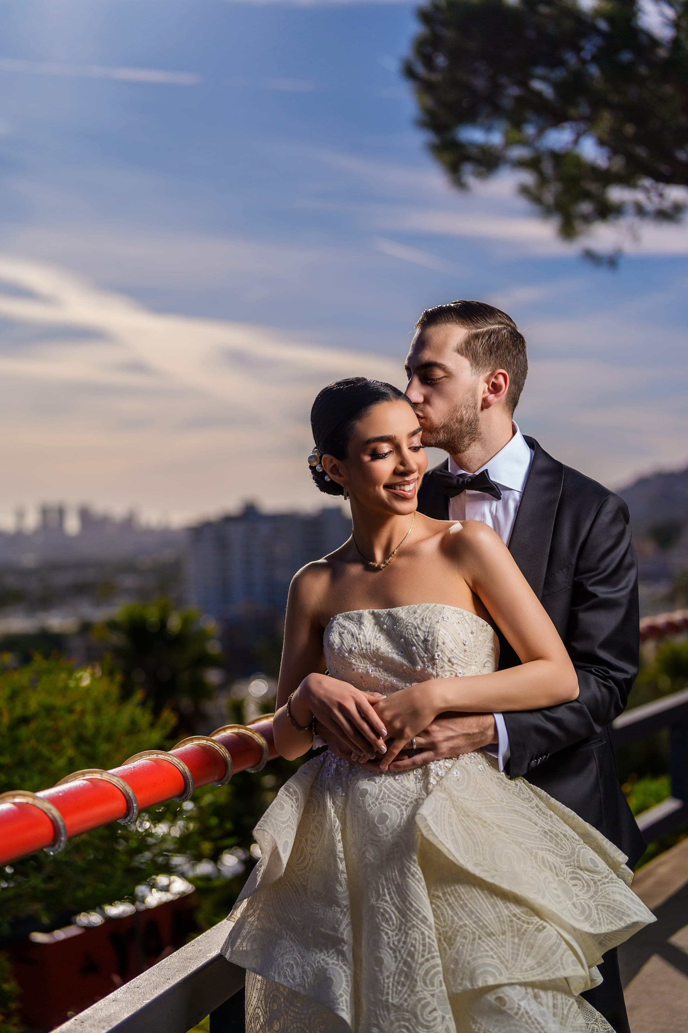 The groom kisses the bride's head at a rooftop railing with the Los Angeles skyline hazy behind them.