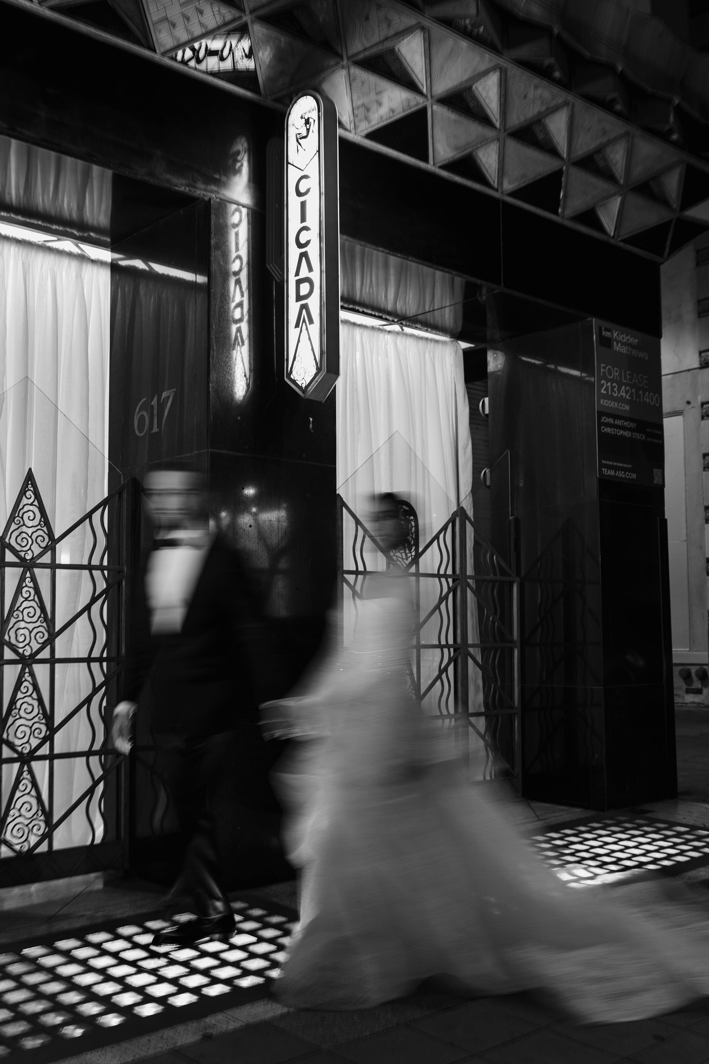 A long exposure blurs the couple walking past Cicada's lighted sign and Art Deco iron gates at night.