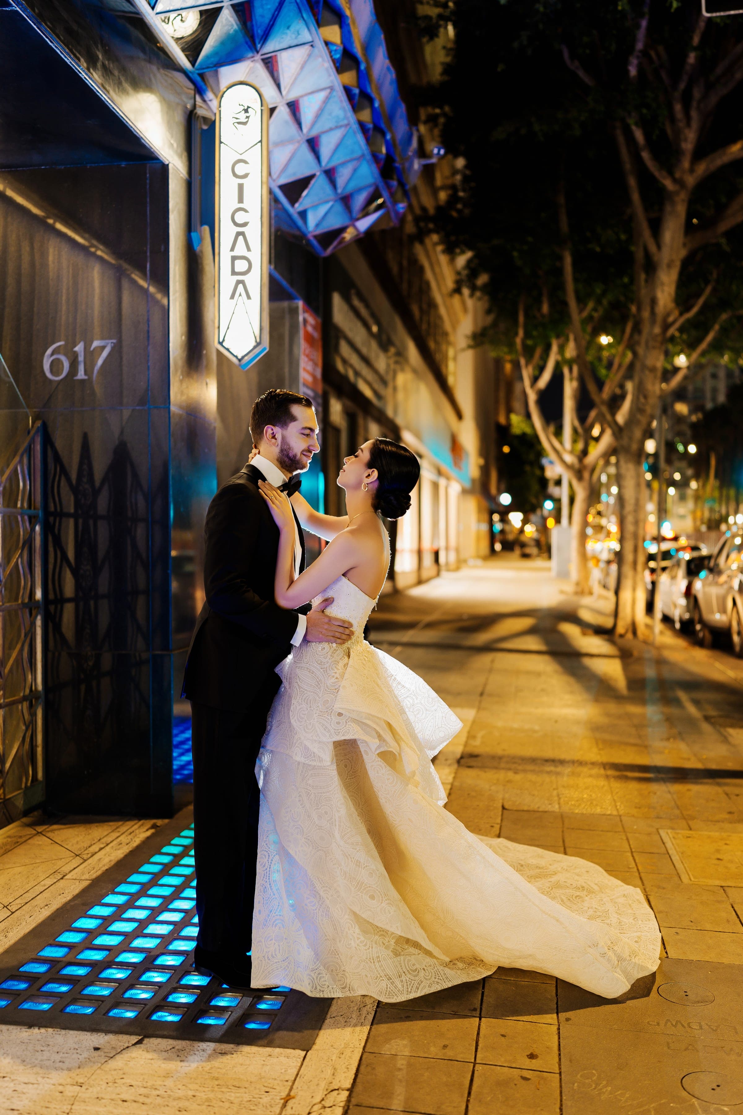 The bride and groom embrace beneath Cicada's glowing Art Deco sign on a Downtown LA sidewalk at night.